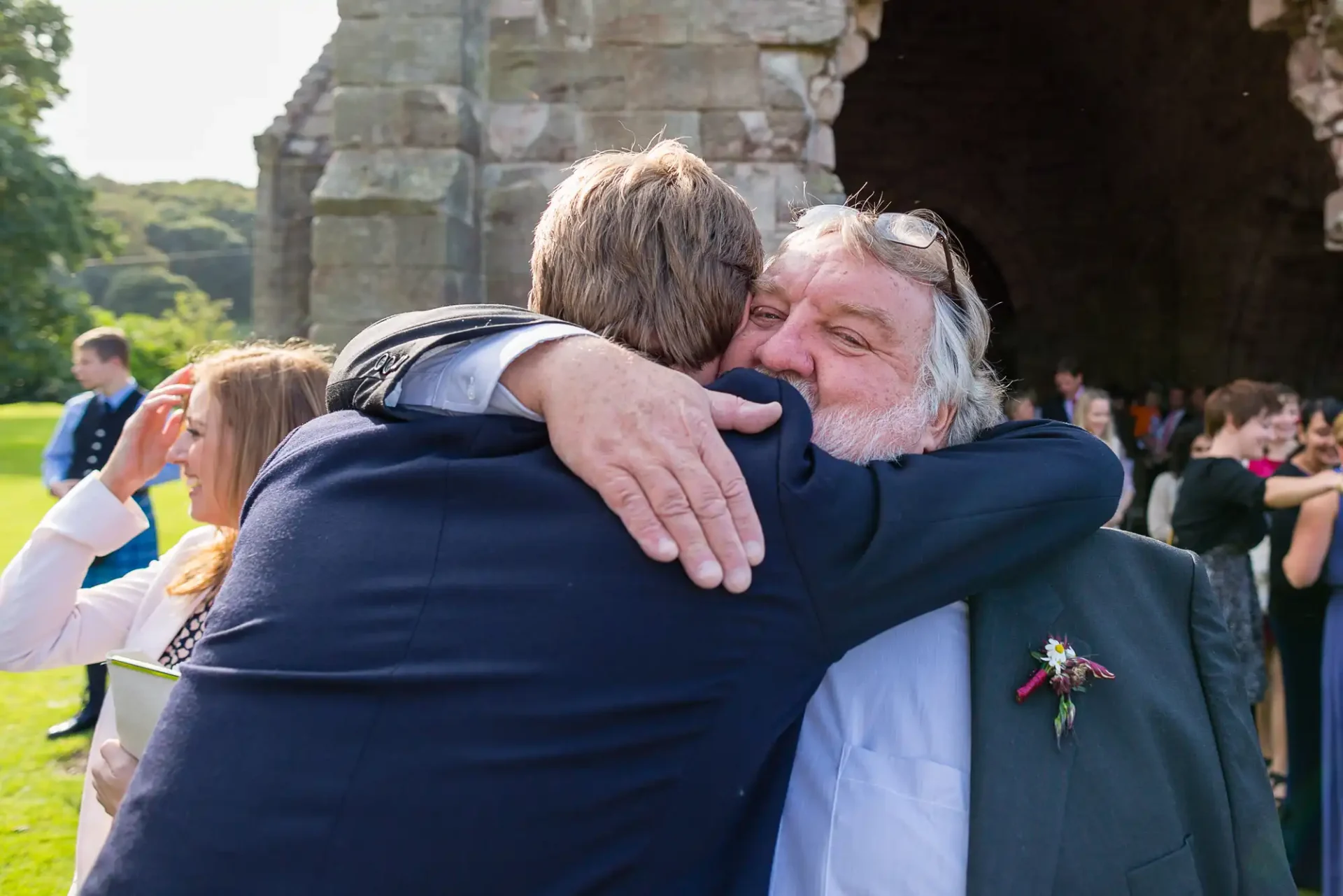 Two men embracing at a sunny outdoor wedding, one with a boutonniere, under an archway with guests in the background.