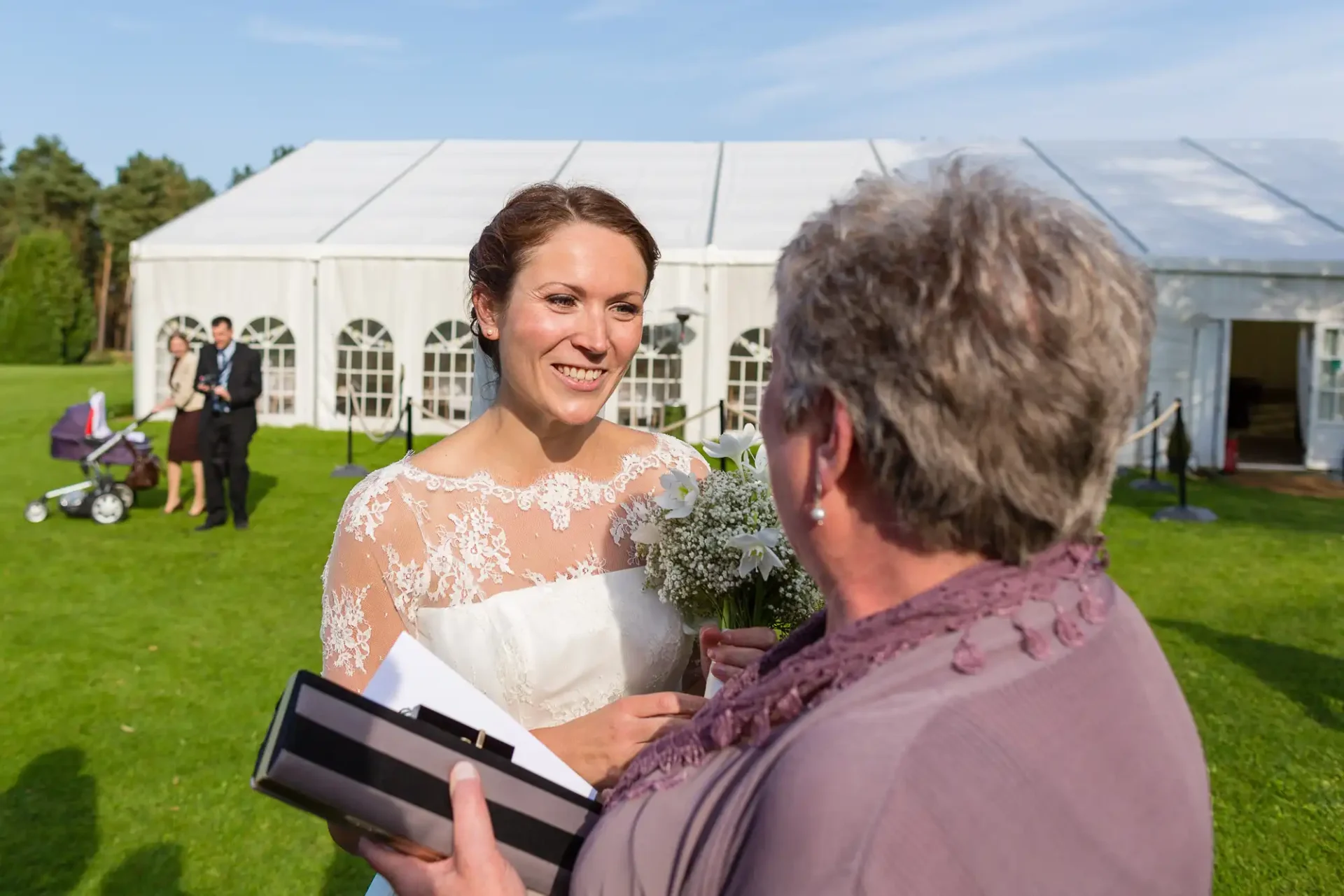 A bride in a lace wedding gown smiling at an older woman holding a gift box, with a wedding marquee and guests in the background on a sunny day.