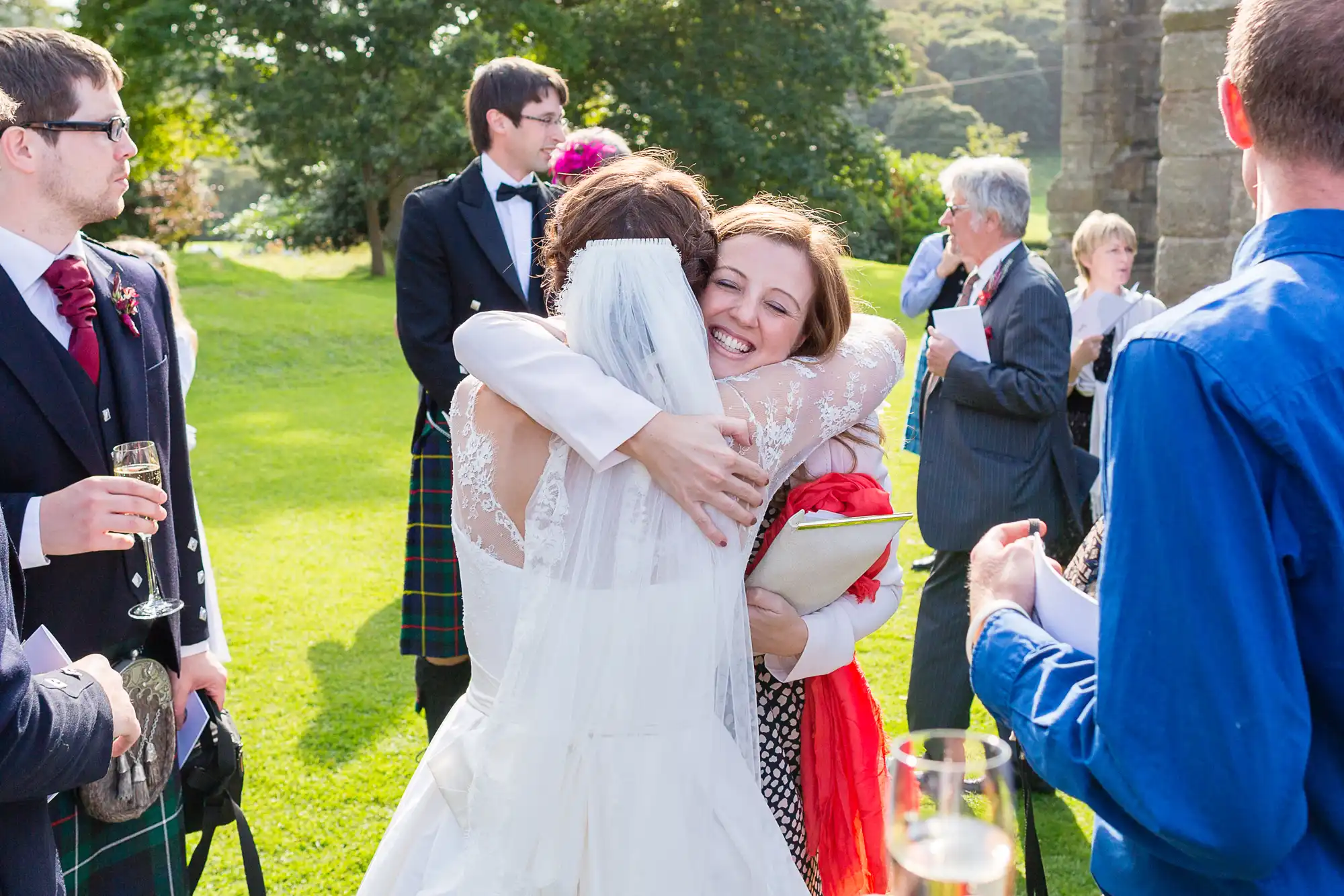 A bride in a white dress hugs a woman in a red shawl at an outdoor wedding ceremony, with guests around them.