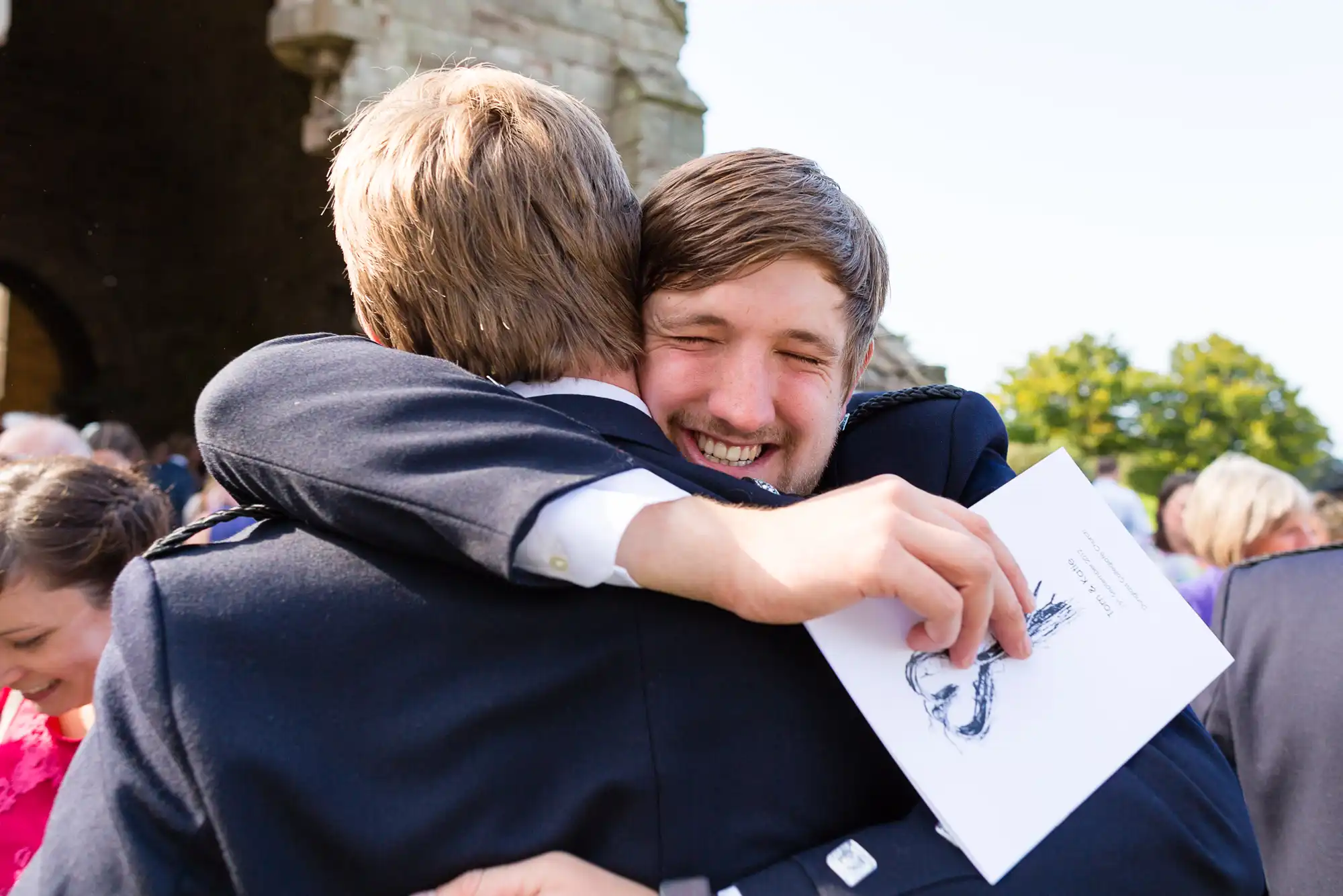 Two men joyfully hugging at an outdoor event, one holding a program with a drawing, both smiling with closed eyes.