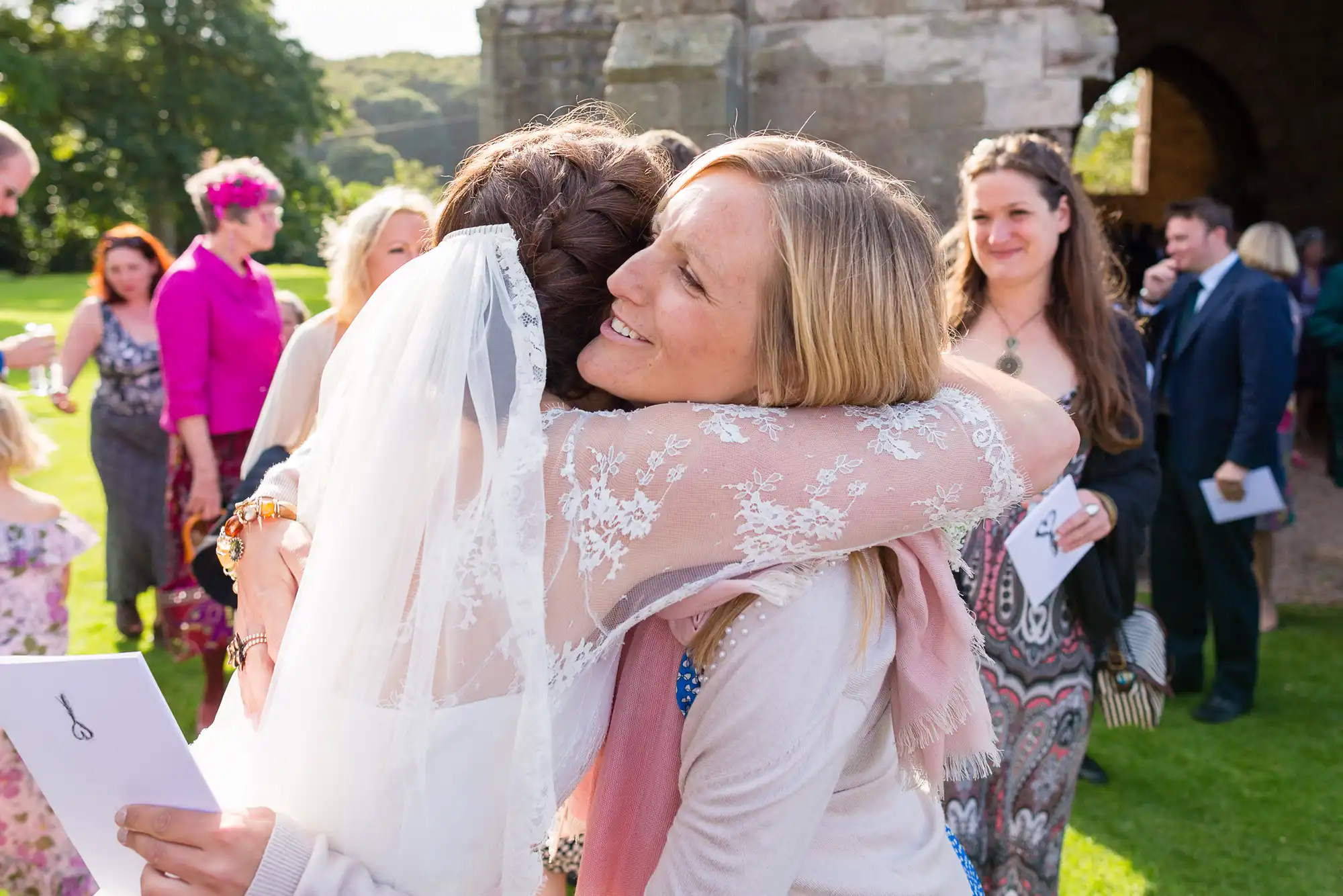 A bride in a lace dress embraces a smiling woman in a blue blouse at a sunny outdoor wedding, with guests in the background.