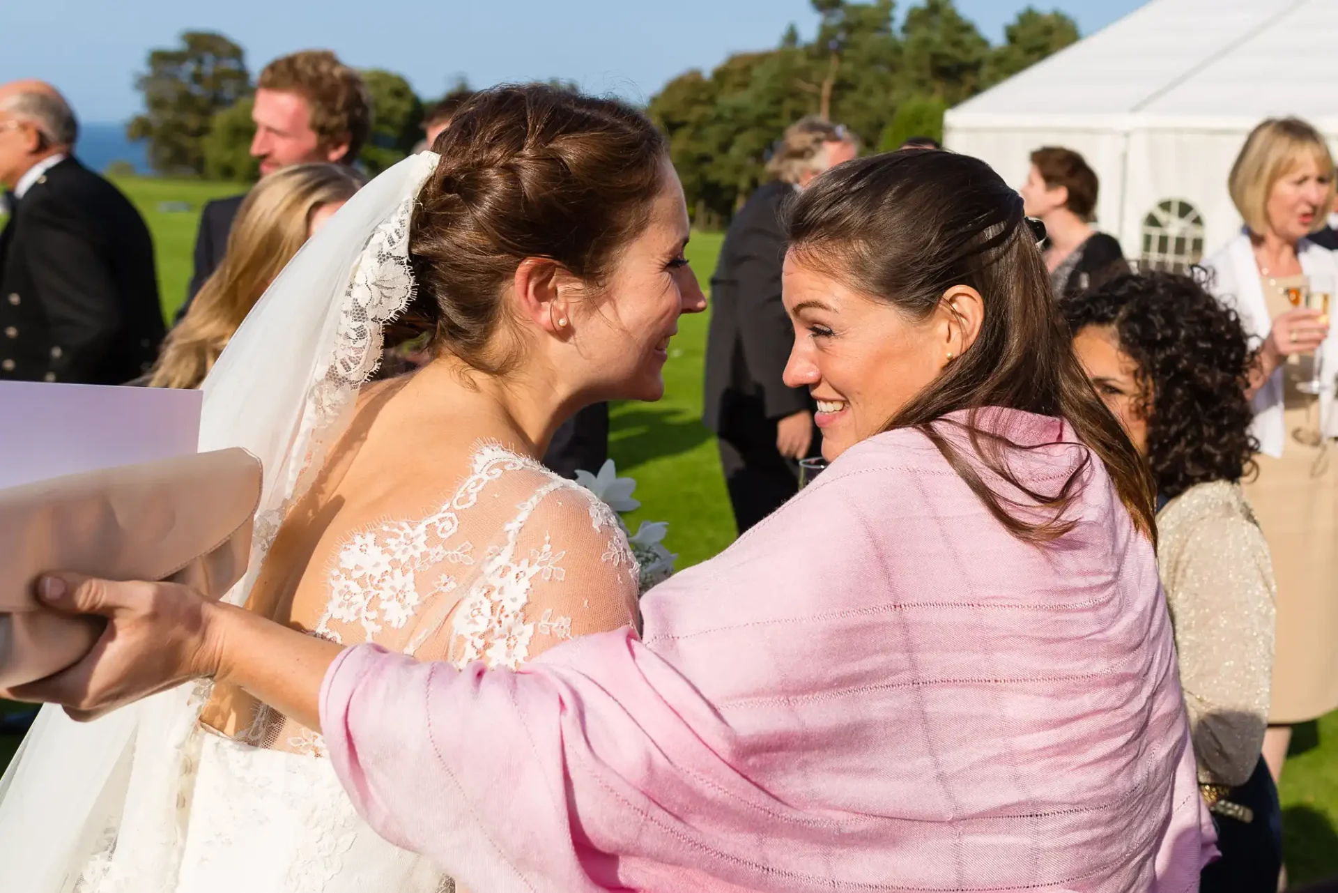 Bride in a lace dress smiling and hugging a woman in a pink jacket at an outdoor wedding reception.
