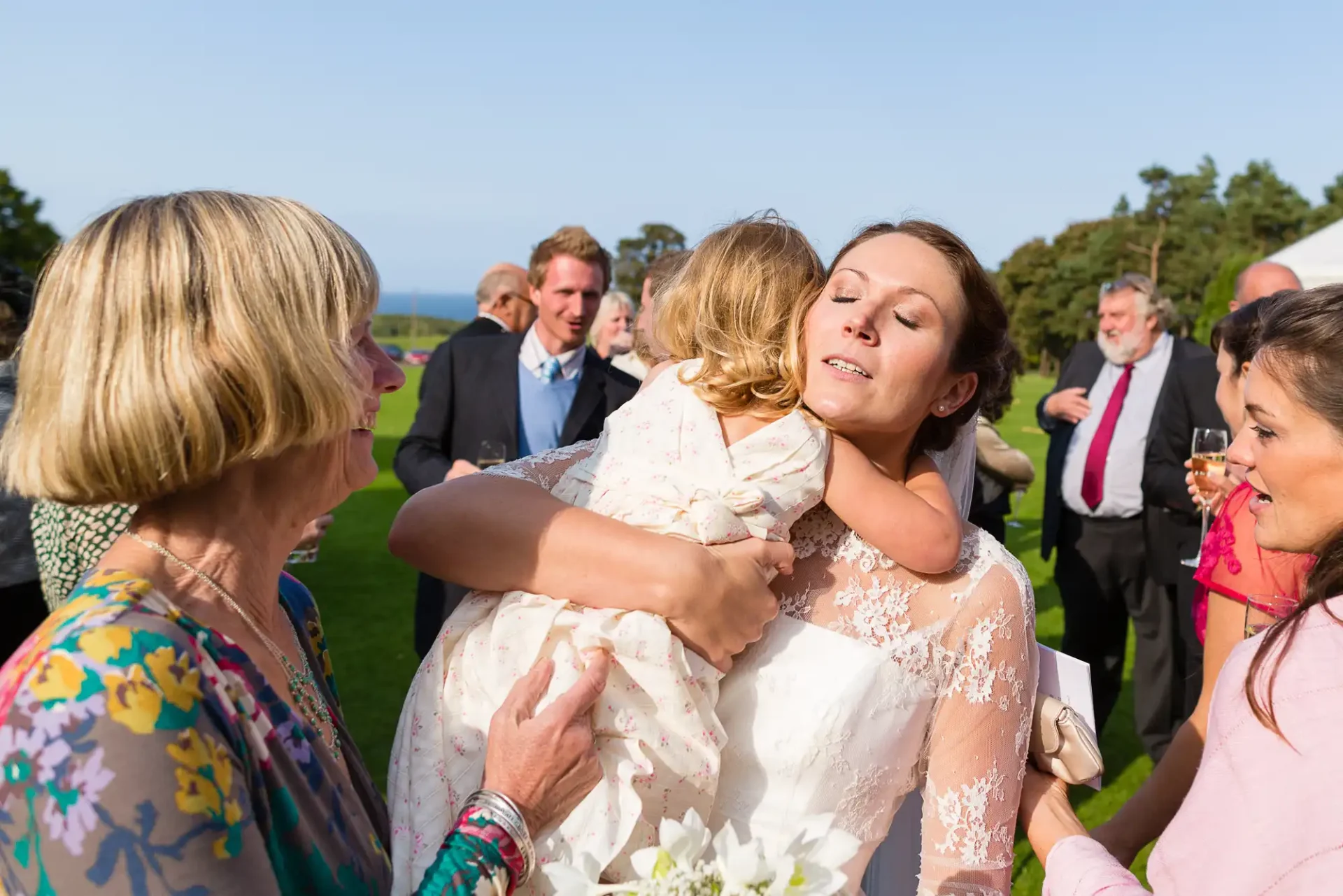 A bride embraces a child while interacting with guests at an outdoor wedding reception.