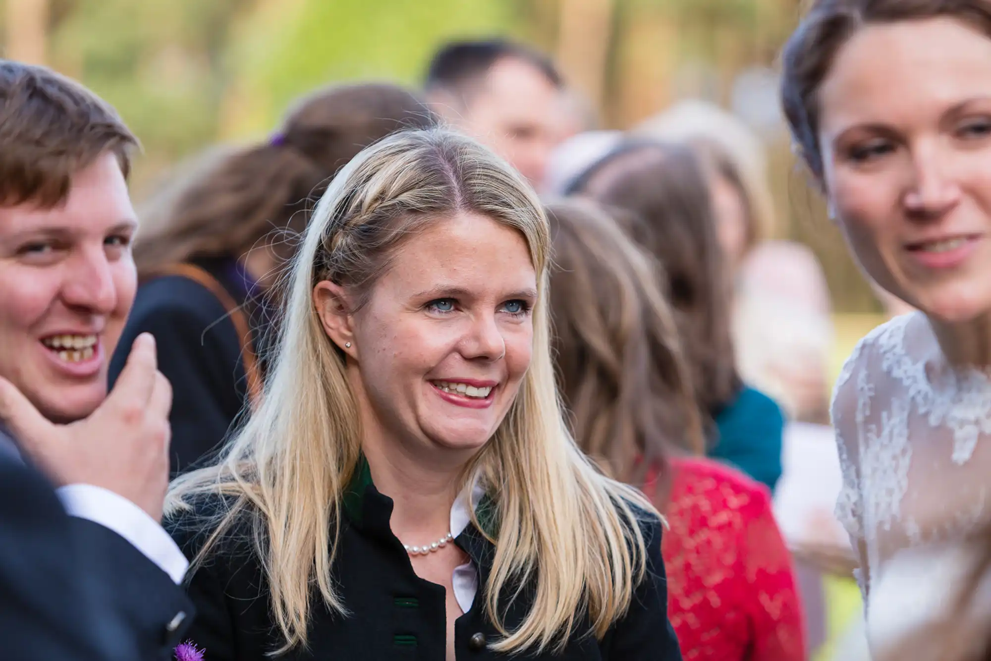 A smiling woman with blond hair attending an outdoor event, surrounded by other guests in partial view.