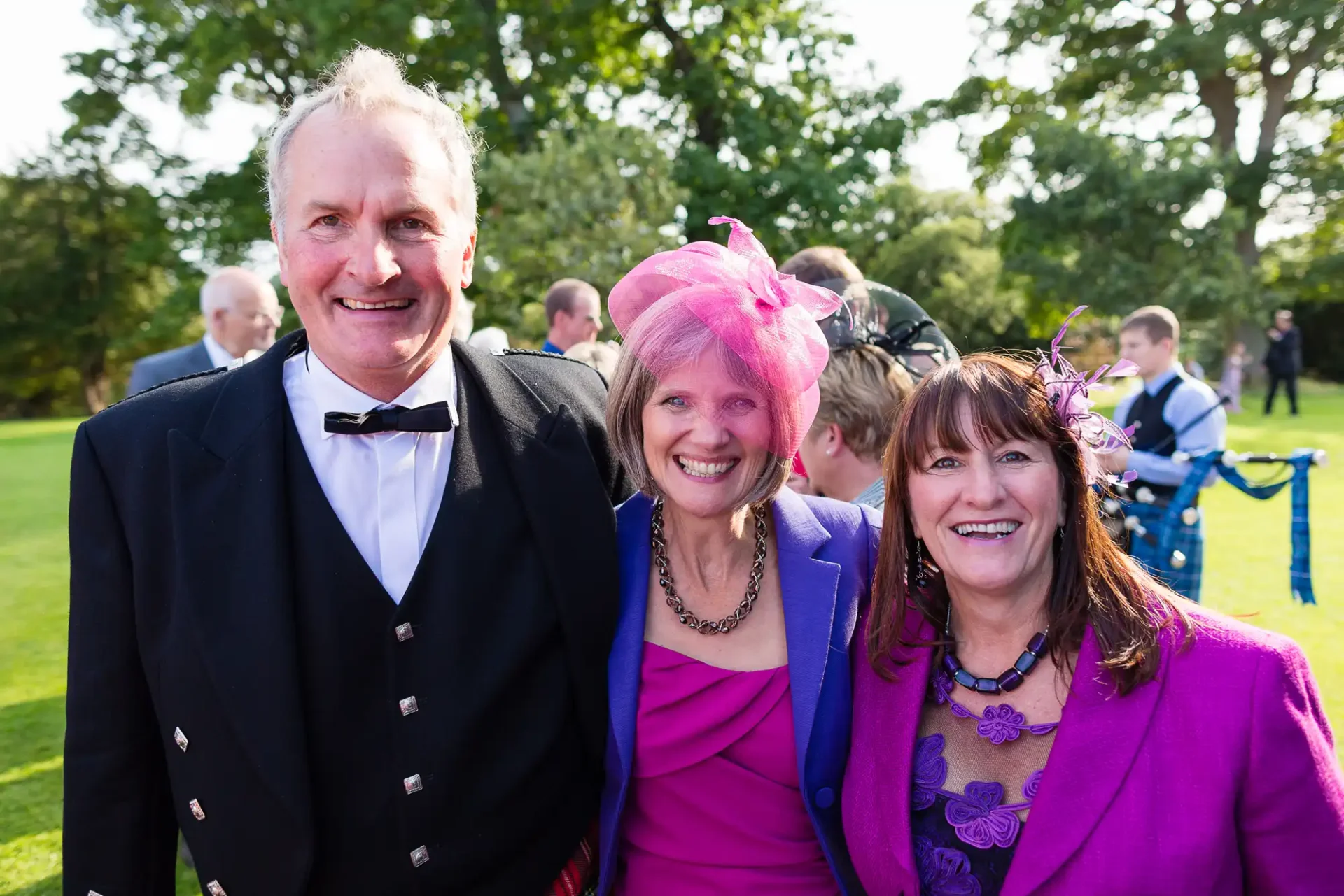 Three smiling adults at an outdoor event; a man in a tuxedo flanked by two women in vibrant purple outfits, one wearing a pink hat.