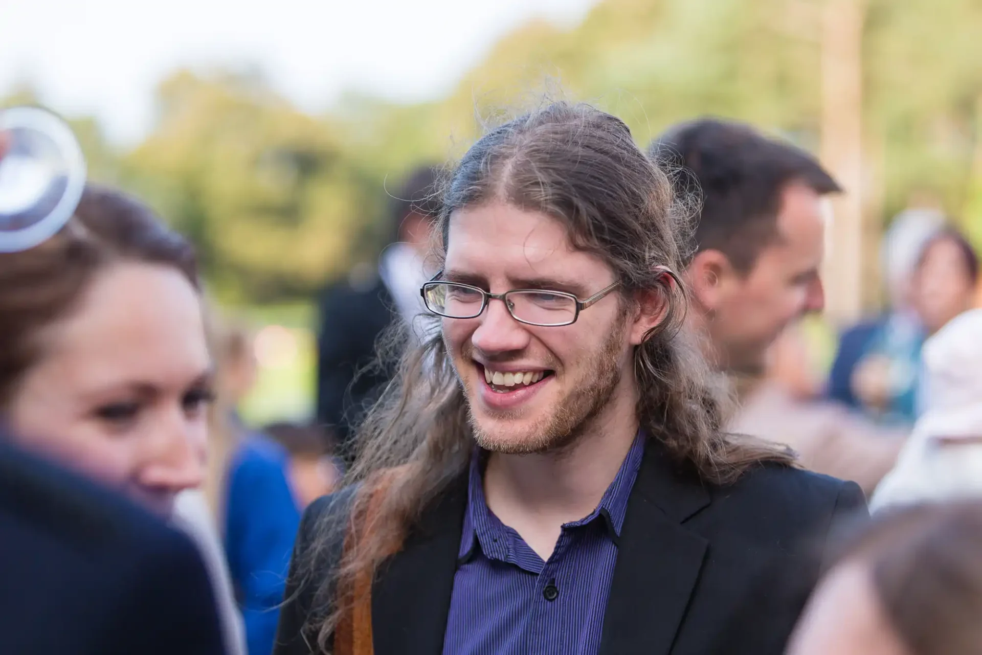 A man with long curly hair and glasses, smiling in a conversation at an outdoor event, dressed in a dark suit.