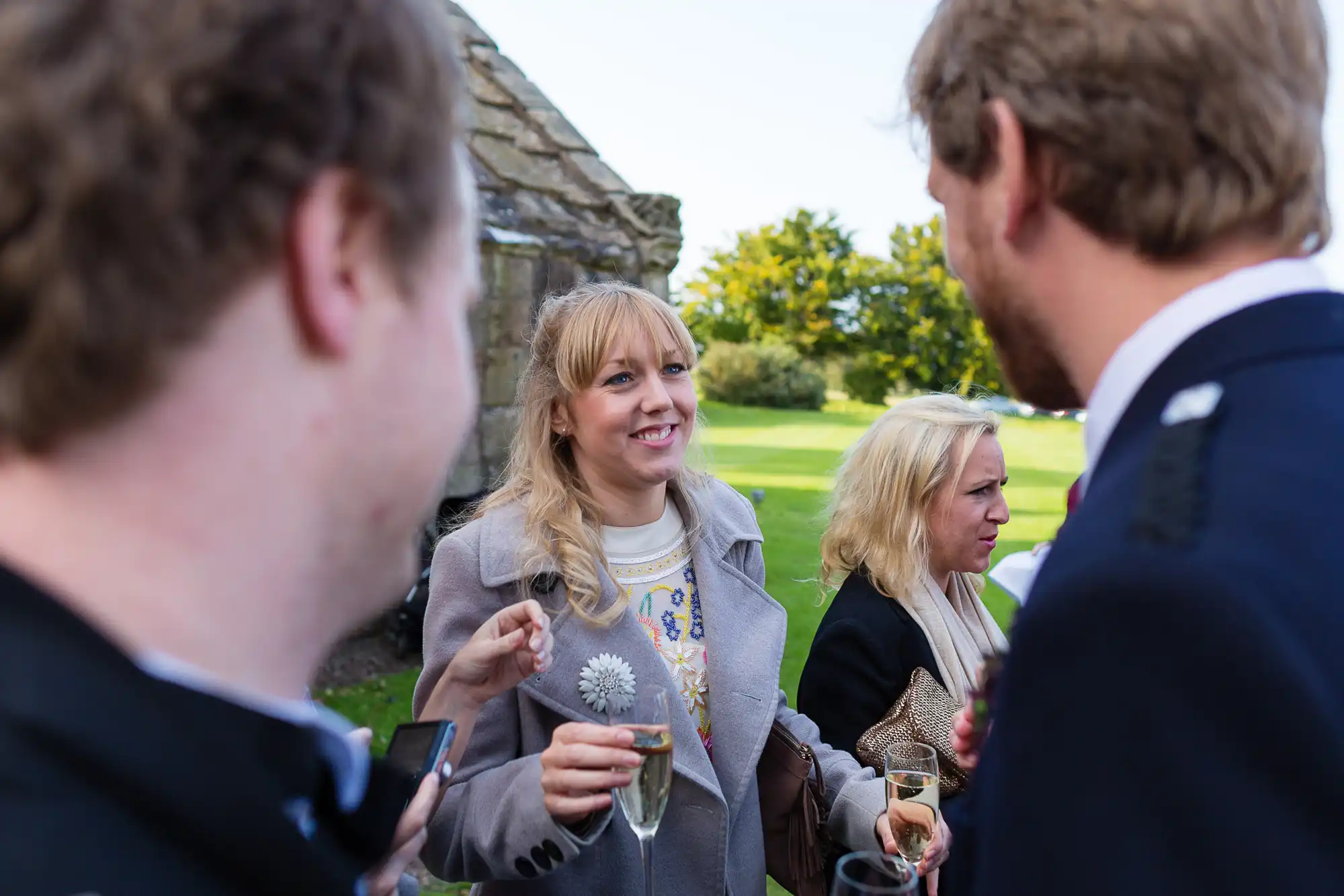 A group of four adults conversing outdoors at a social event, one woman holding a wine glass and a dandelion.