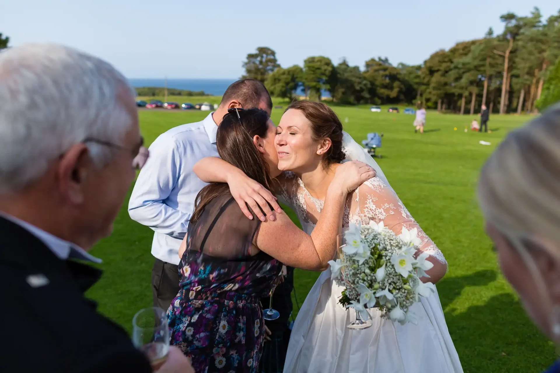 A bride in a white dress hugs a woman in a floral dress, receiving a kiss on the cheek, with guests around them at an outdoor wedding setting.