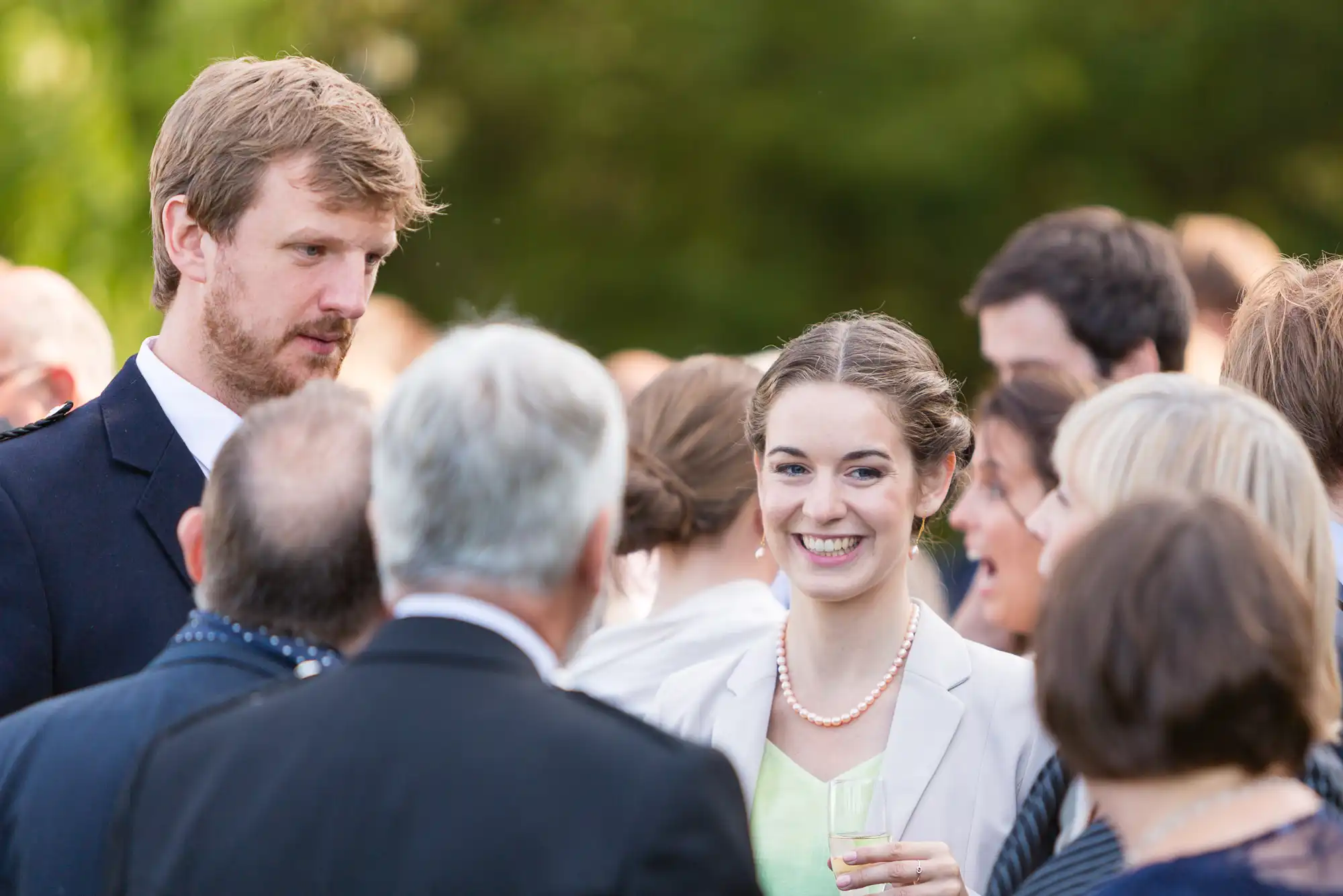 A young woman in a white jacket smiling while holding a drink at an outdoor social gathering, surrounded by other guests conversing.