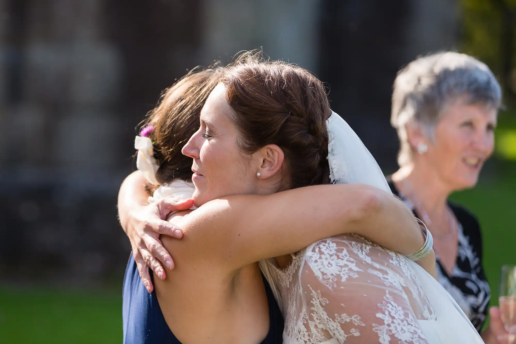 Bride in a lace dress embracing a bridesmaid with an older woman smiling in the background at an outdoor wedding.