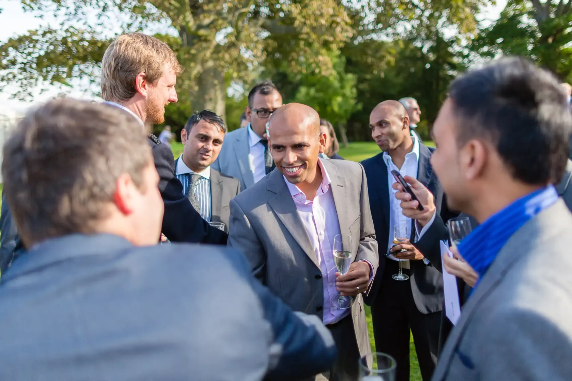 Group of men in business attire conversing at an outdoor event, some holding drinks. one man in the center is smiling broadly.