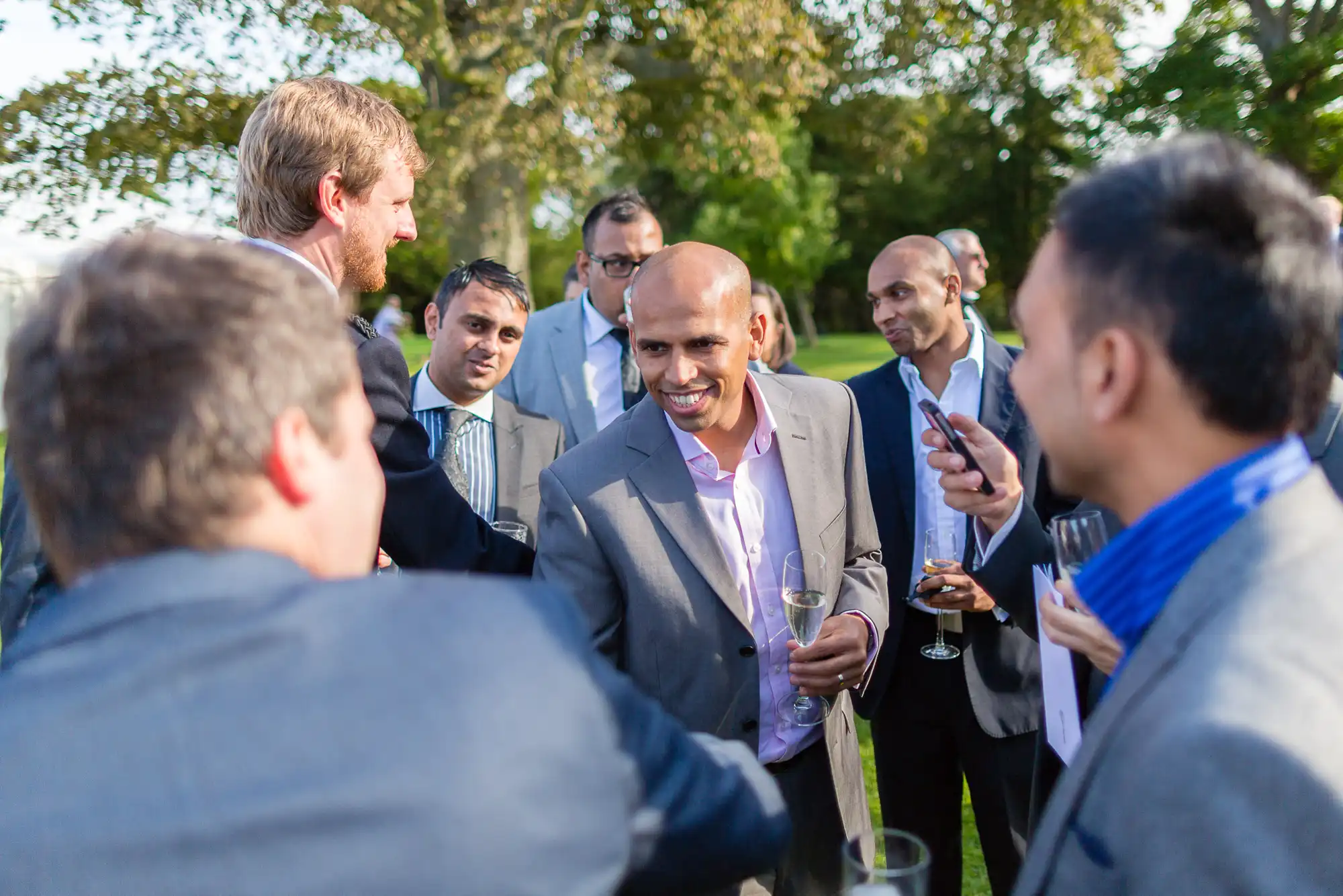 Group of men in business attire conversing at an outdoor event, some holding drinks. one man in the center is smiling broadly.