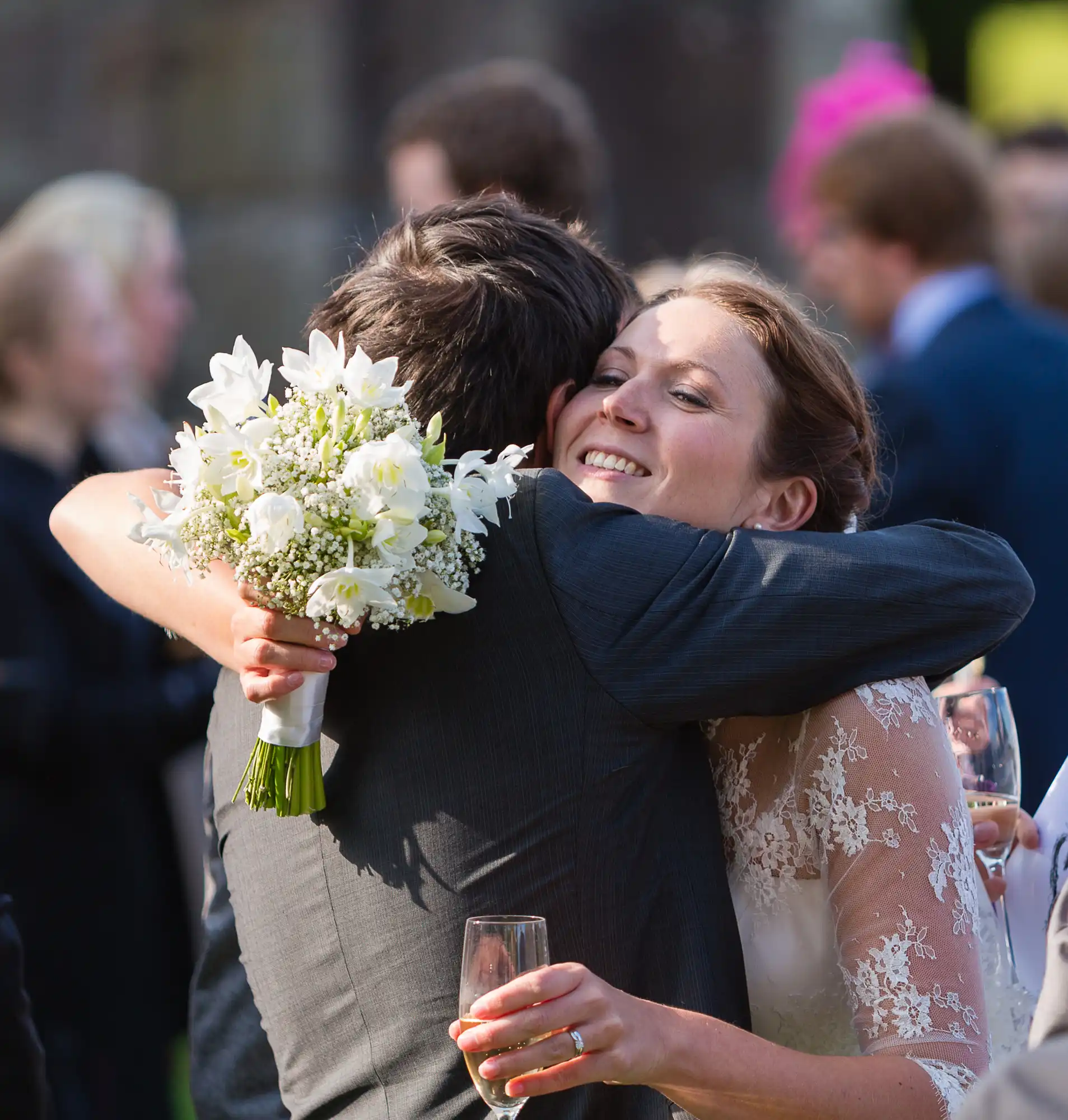 Bride in lace dress hugging a guest at a wedding, holding a bouquet of white flowers, with other guests in the background.