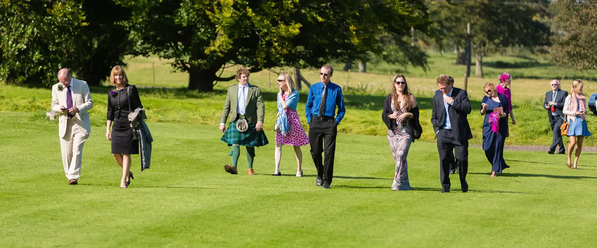 A group of elegantly dressed adults walks across a green lawn, each carrying different accessories, under a clear sky.