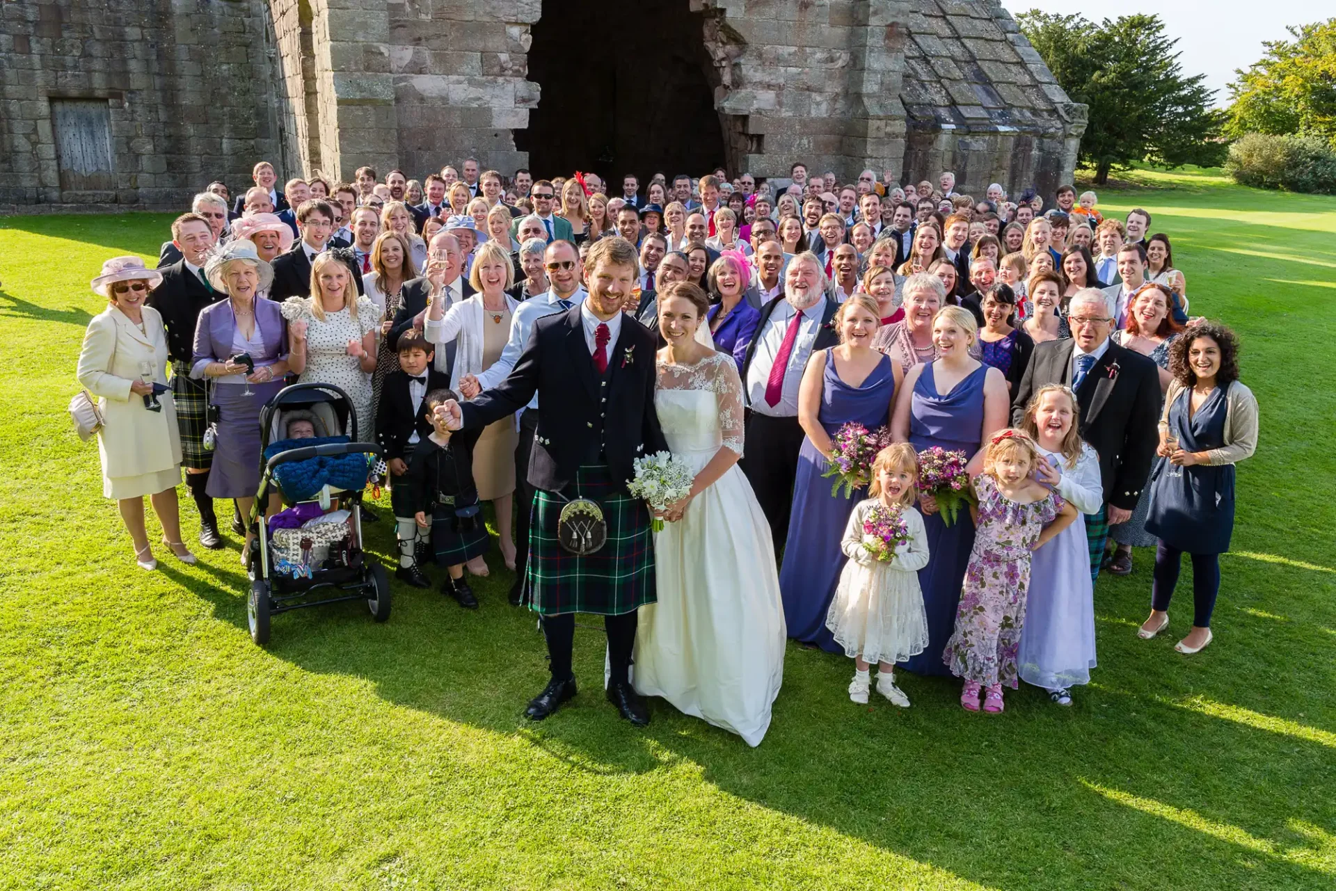 A large wedding group photo on a sunny day, featuring a bride and groom in the center, surrounded by guests in formal attire.