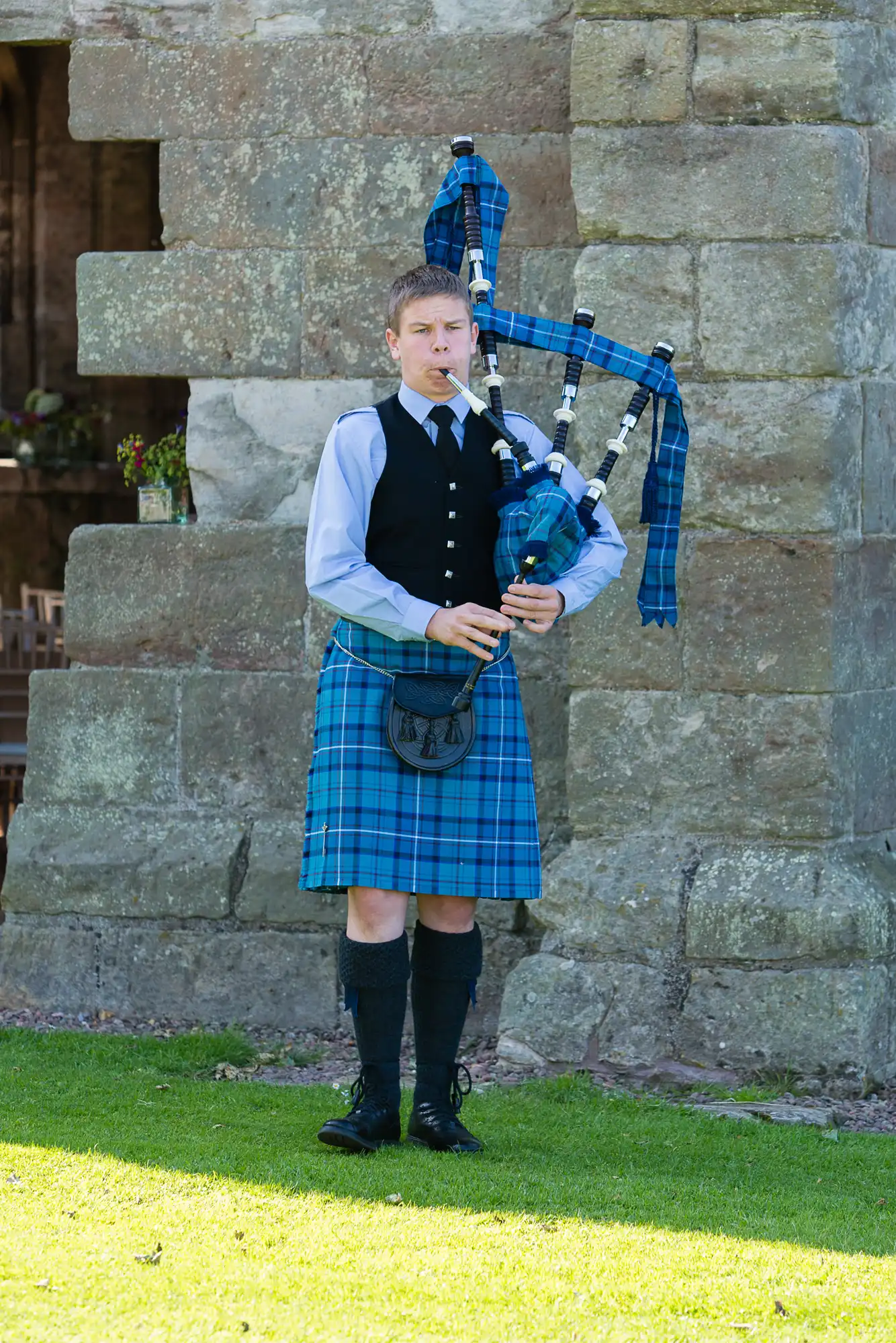 A young man in traditional scottish attire, including a kilt and sporran, playing the bagpipes outdoors by a stone wall.
