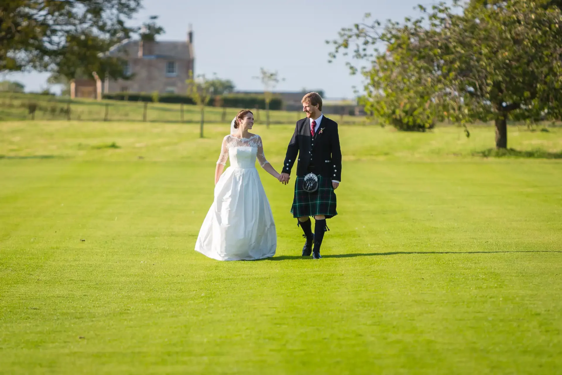A bride in a white gown and a groom in a kilt walking hand in hand across a sunlit grassy field with a house in the background.