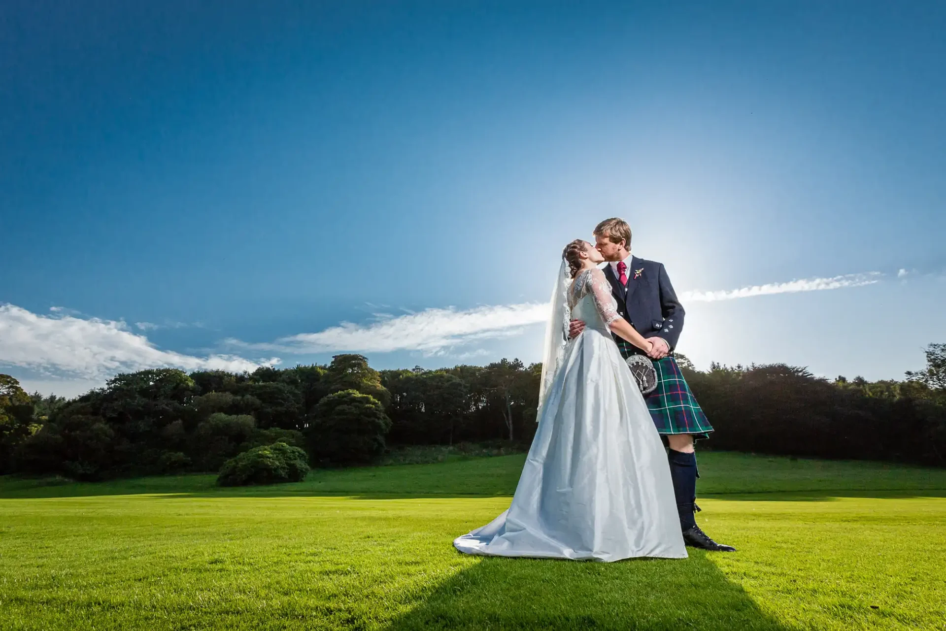 A bride and groom embracing on a sunny grassy field, the groom in a kilt and the bride in a long white gown, with a clear blue sky above.