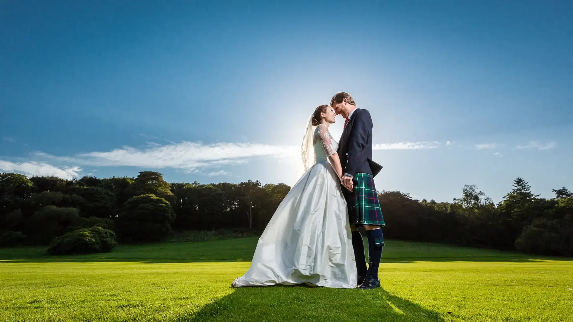 A bride and groom in a sunlit field, the bride in a white gown and the groom in a kilt, share a kiss under a bright sky.
