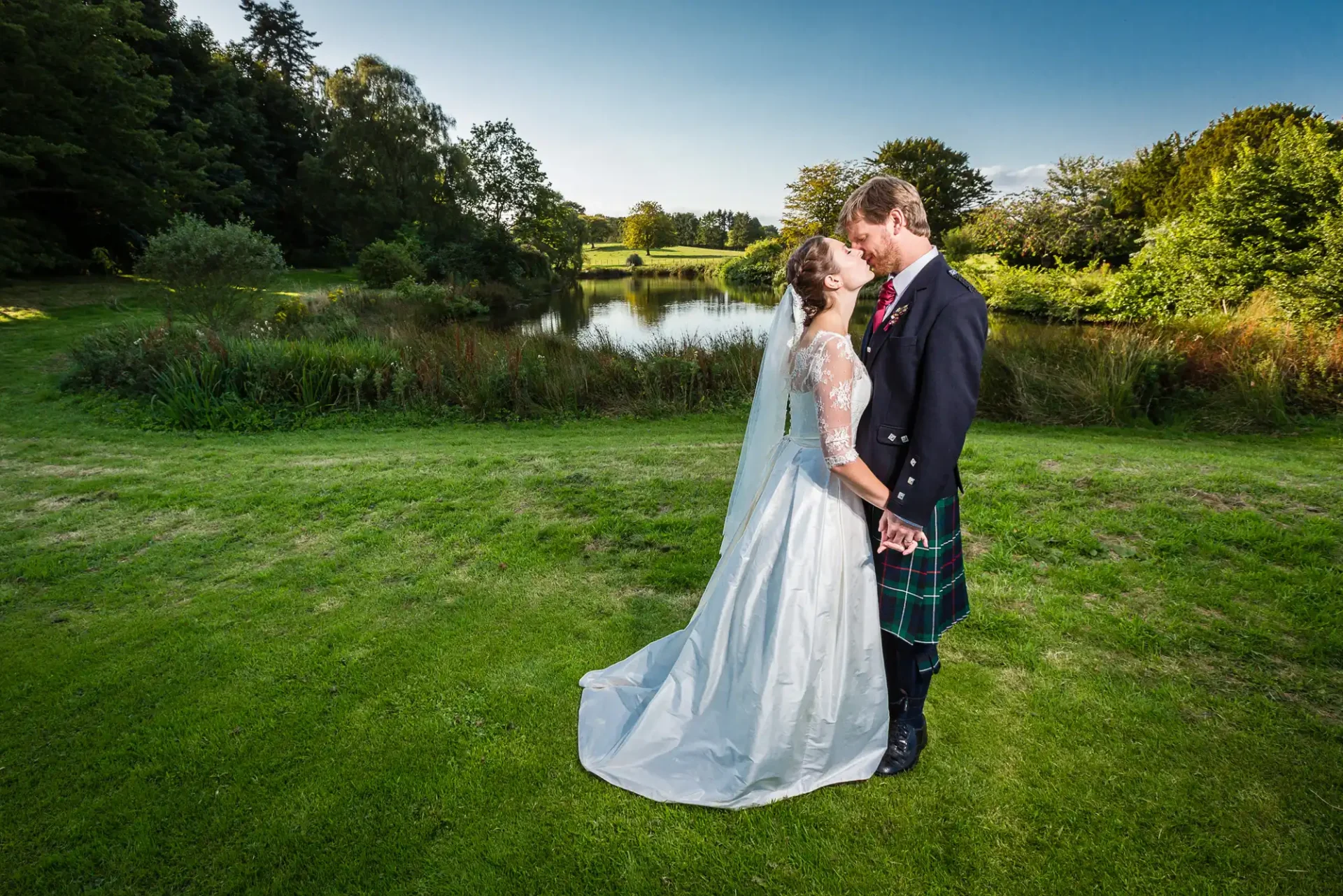 A newlywed couple embraces on a grassy field beside a pond, with the groom in a kilt and the bride in a white gown, surrounded by lush greenery under a clear sky.