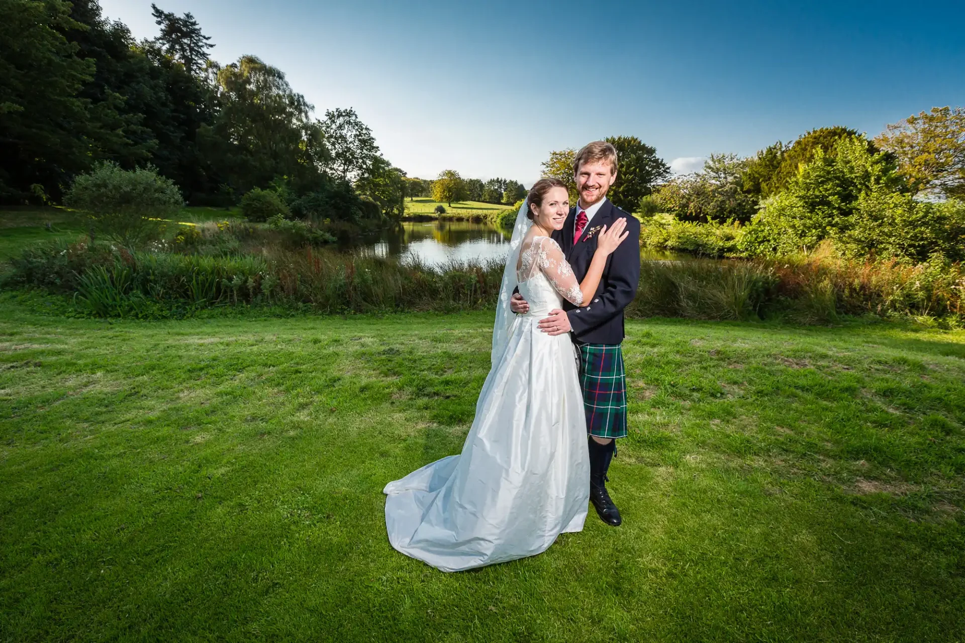 A bride in a white gown and a groom in a kilt embracing on a grassy field with a pond and trees in the background.