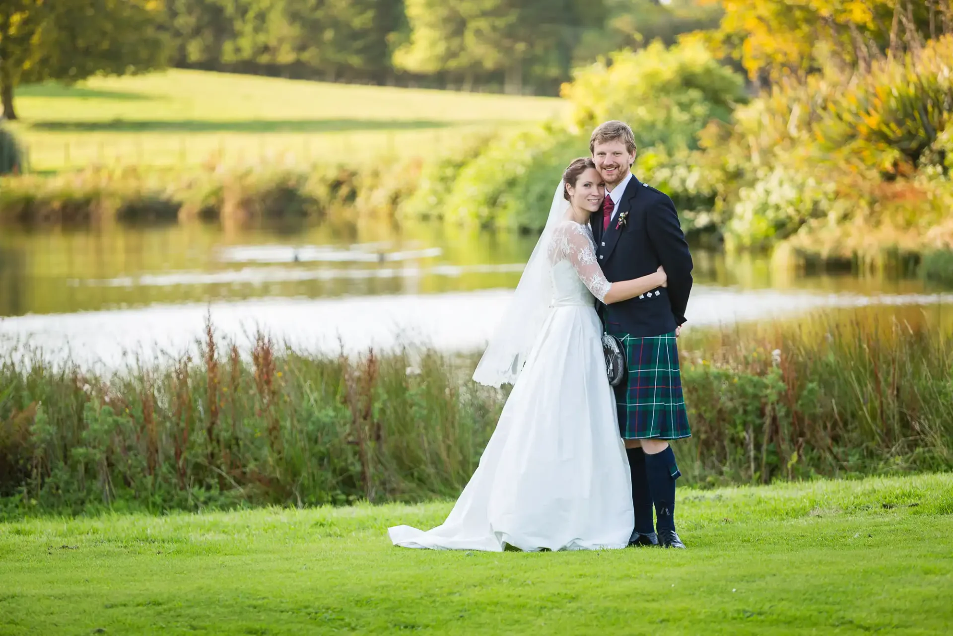 A bride in a white dress and a groom in a kilt embracing near a pond with greenery in the background.
