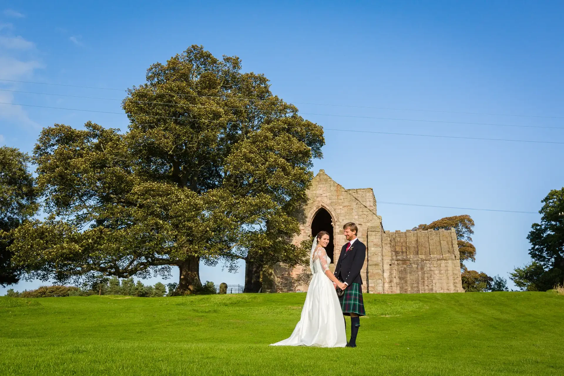 Bride in a white dress and groom in a kilt standing by a ruin under a large tree in a sunny, grassy field.