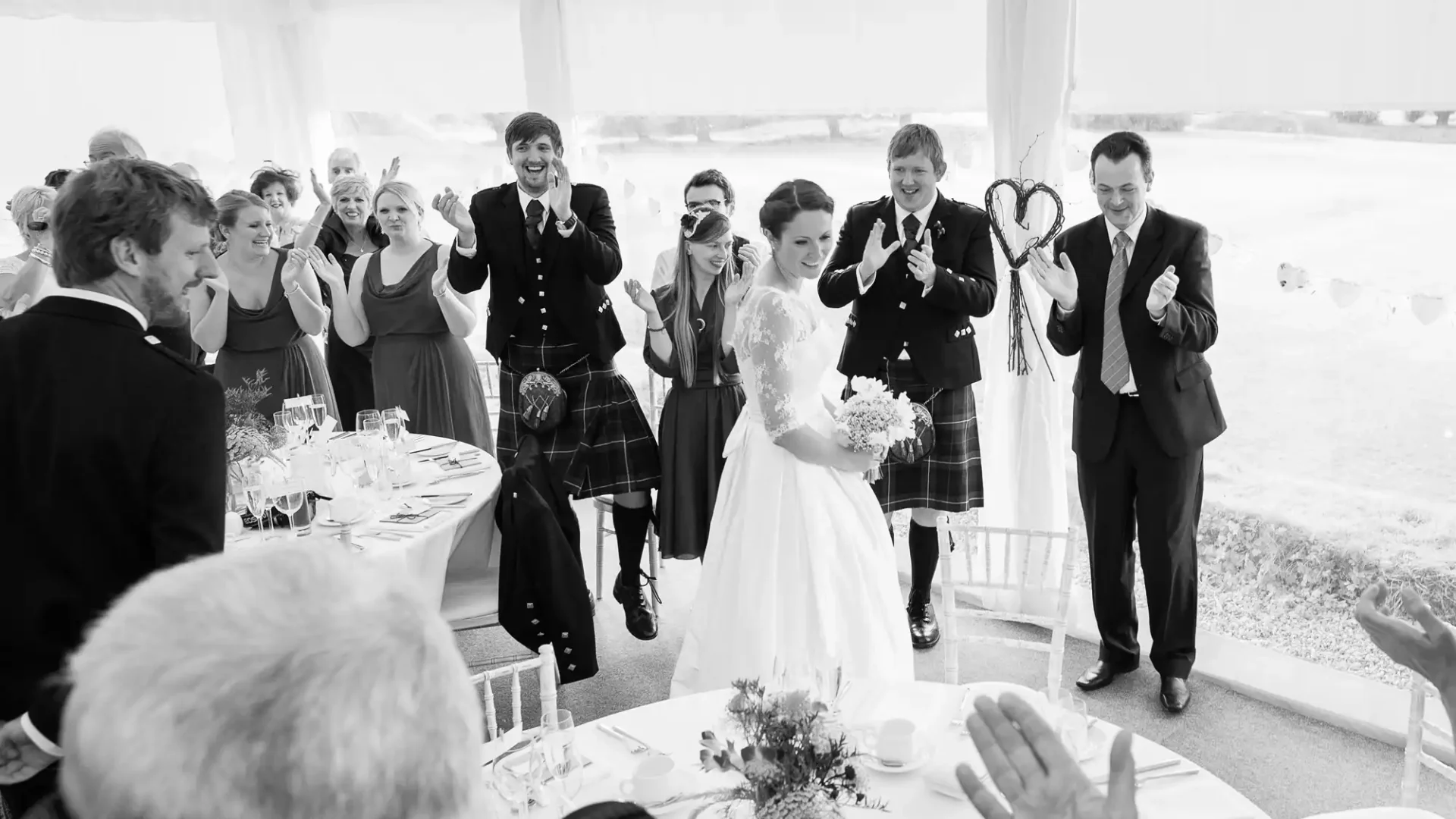 Bride and groom walking hand-in-hand through a clapping crowd at their wedding reception, guests wearing traditional scottish attire.