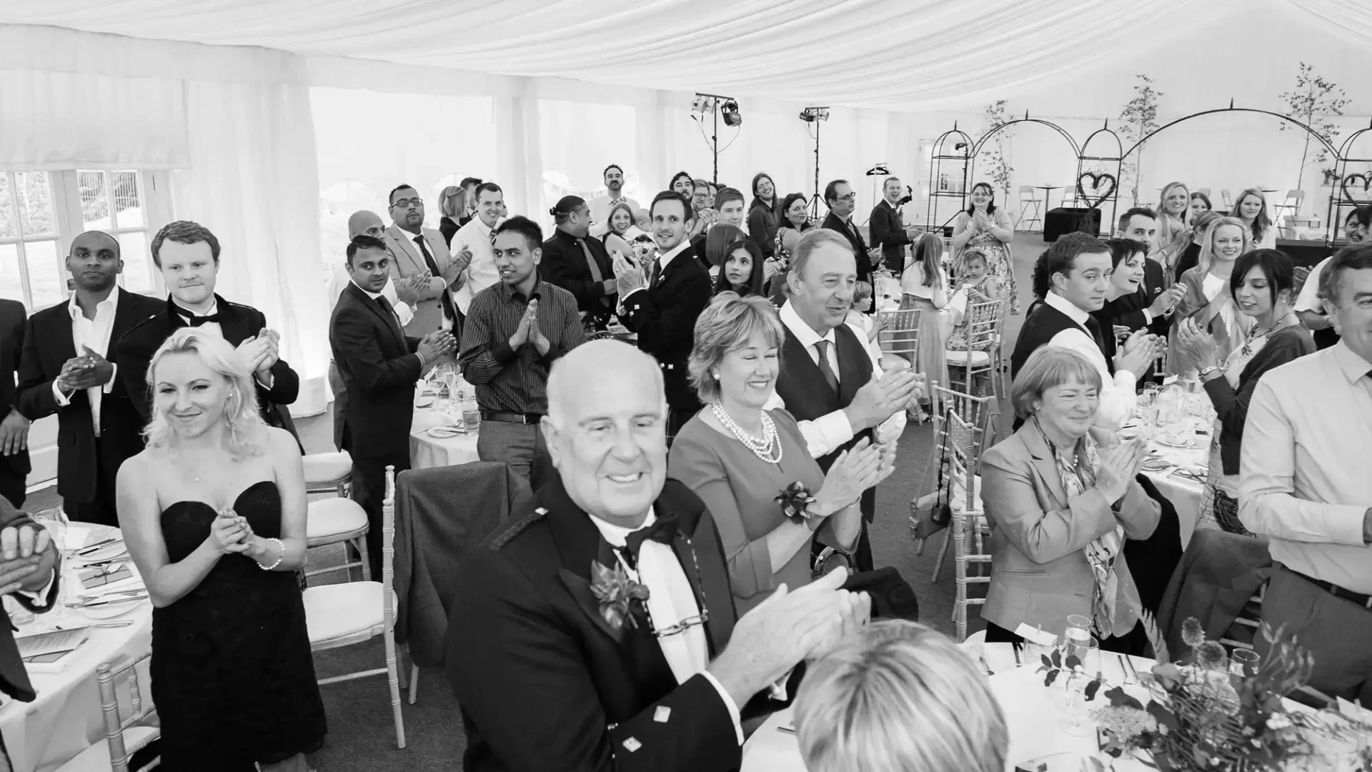 Guests in formal attire applauding at a wedding reception in a tent, image in black and white.
