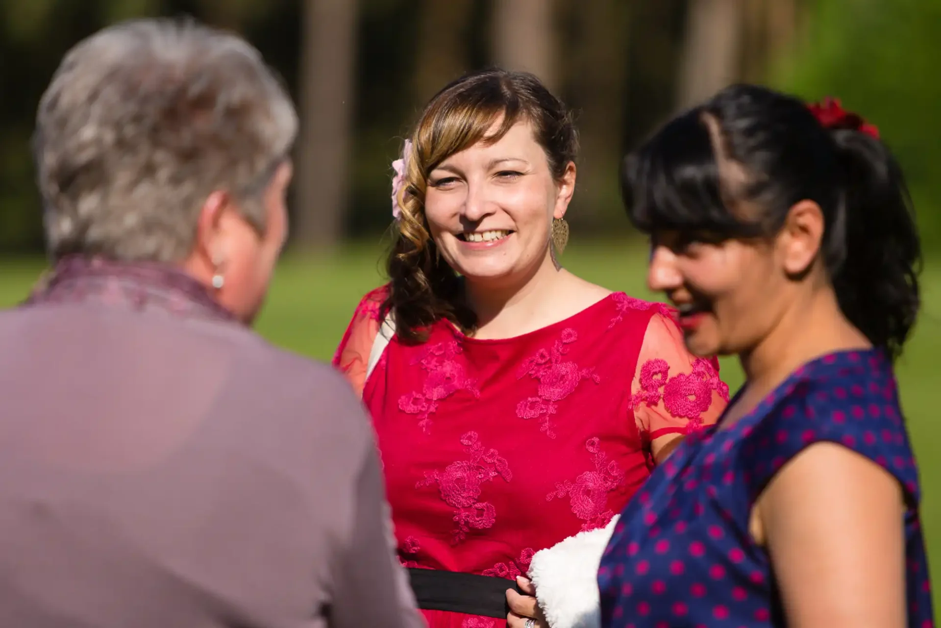 Two women in red dresses smiling and conversing with another person outdoors on a sunny day.