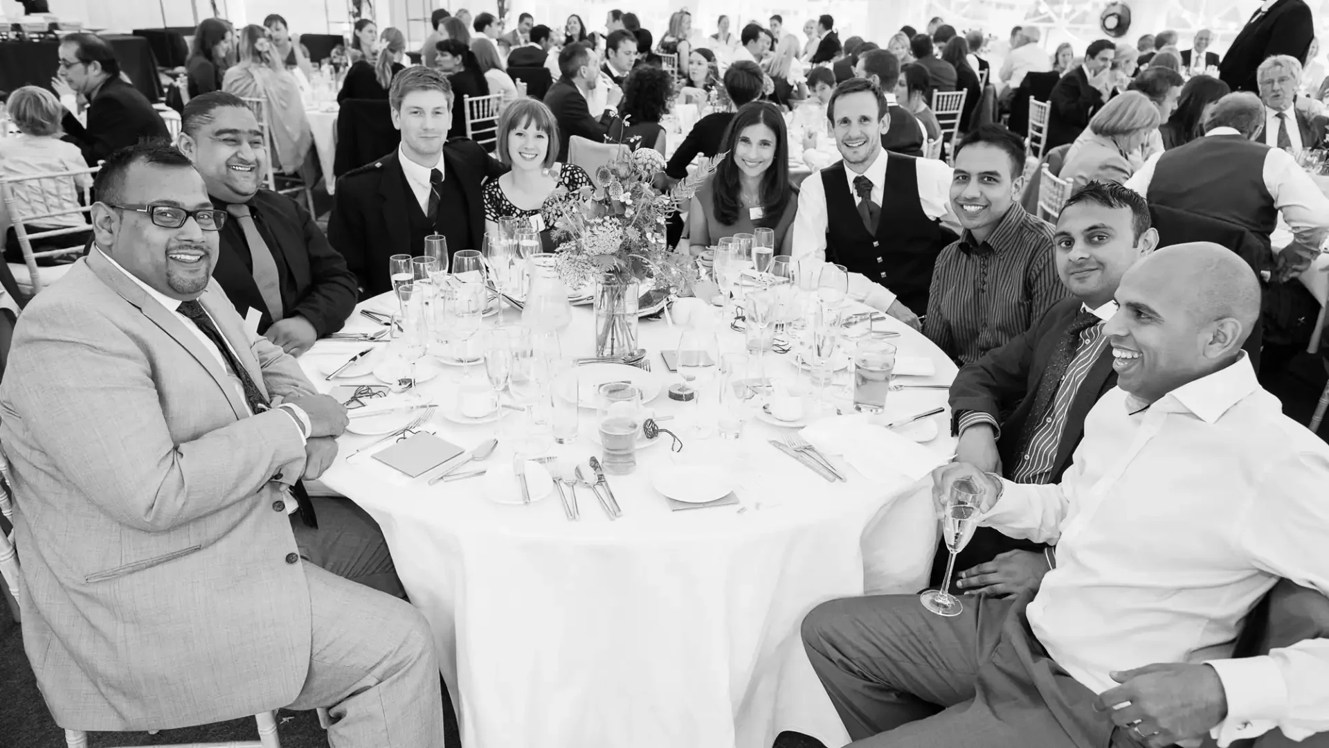 A group of people smiling at a table during a formal event in a tent, dressed in business attire. black and white photograph.