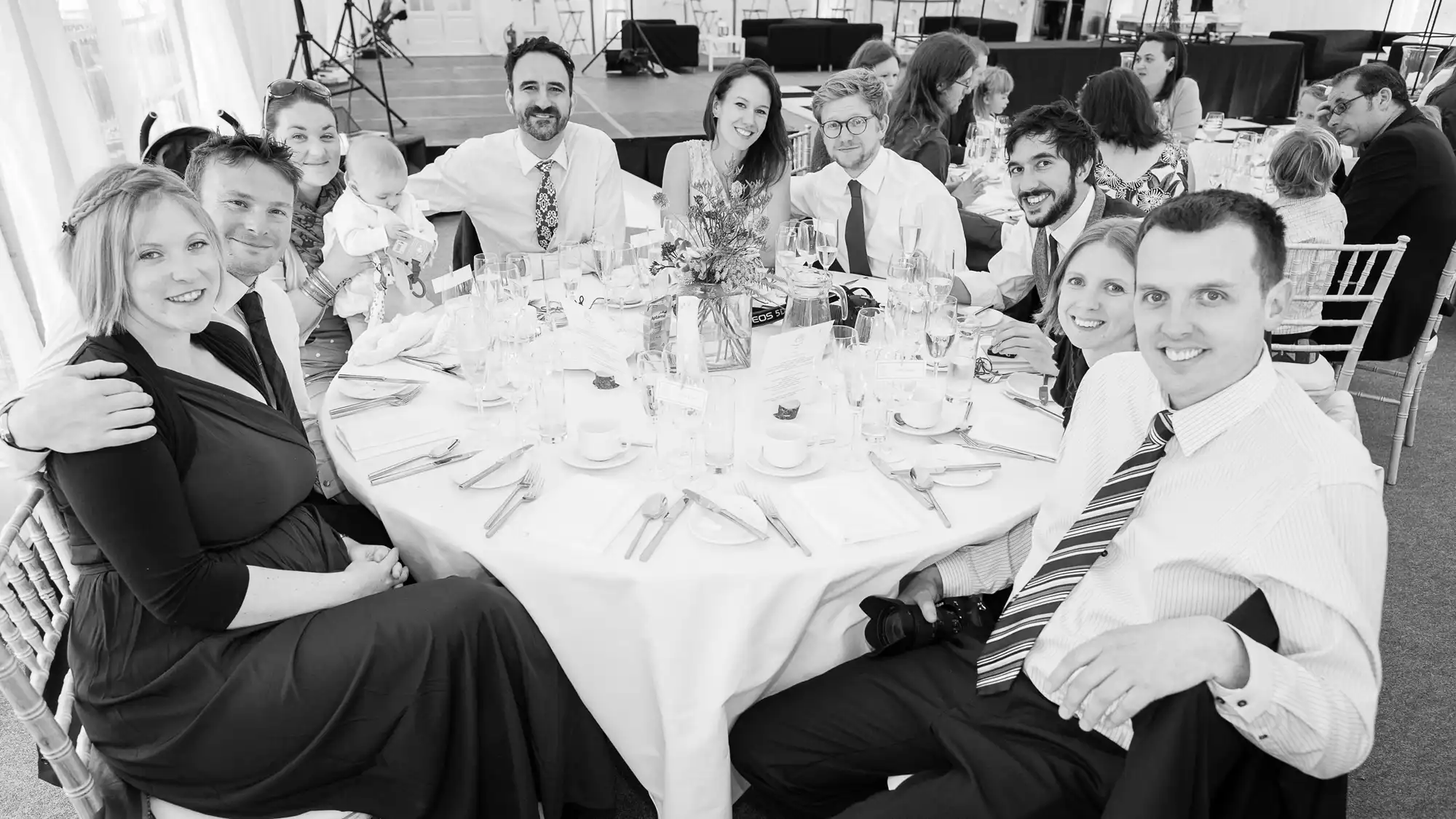 Group of adults smiling at a table during a wedding reception, some holding babies, in a black and white photo.