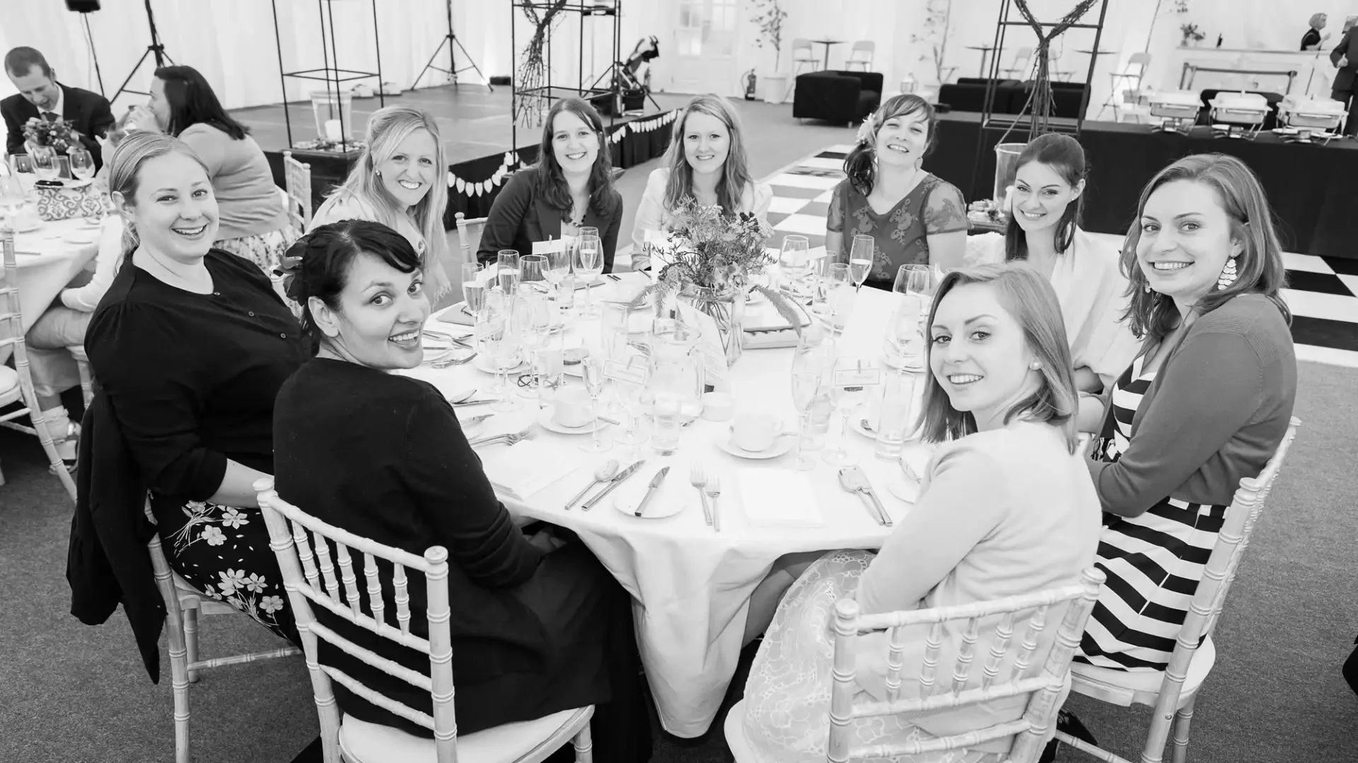 A group of nine women smiling at a formal dining event in a large tent, seated around a table set with dinnerware and glasses. the image is in black and white.