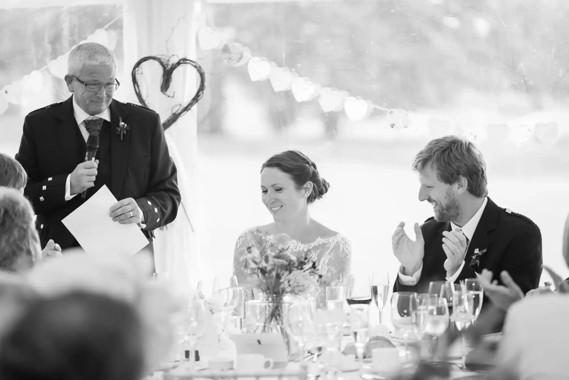 A black and white photo of a wedding reception where an older man is giving a speech, and a laughing couple is listening, seated at a decorated table.