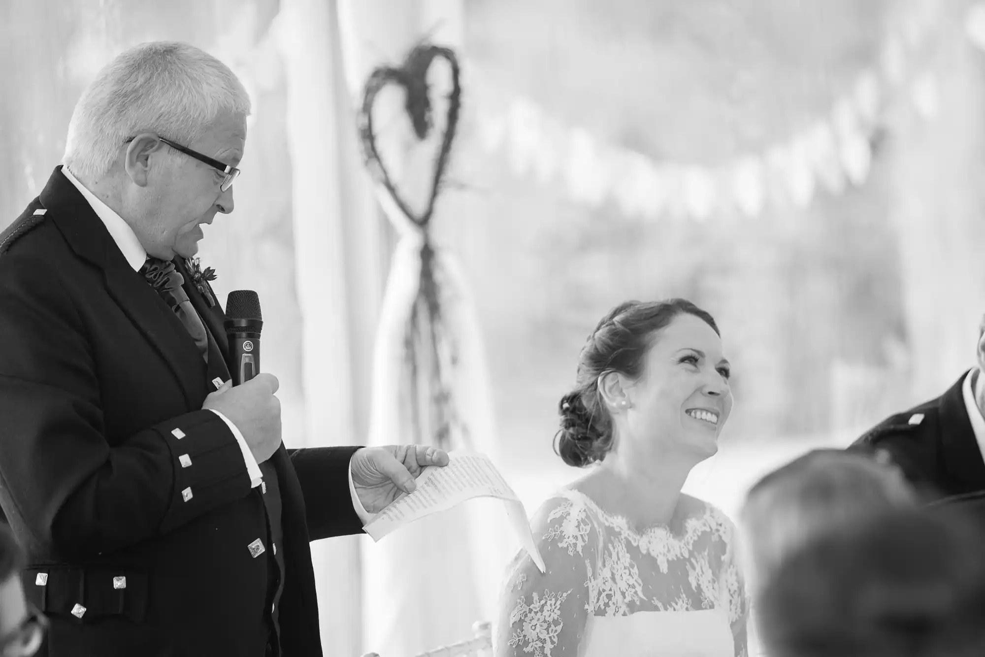 A man in a suit reads from a paper, speaking into a microphone at a wedding reception, as a smiling bride in a lace dress listens.