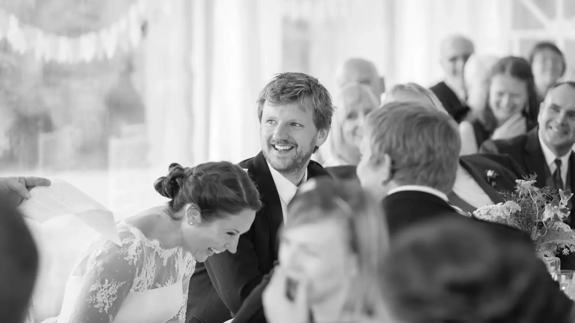 A black and white photo of a smiling bride and groom at a wedding reception, surrounded by guests.