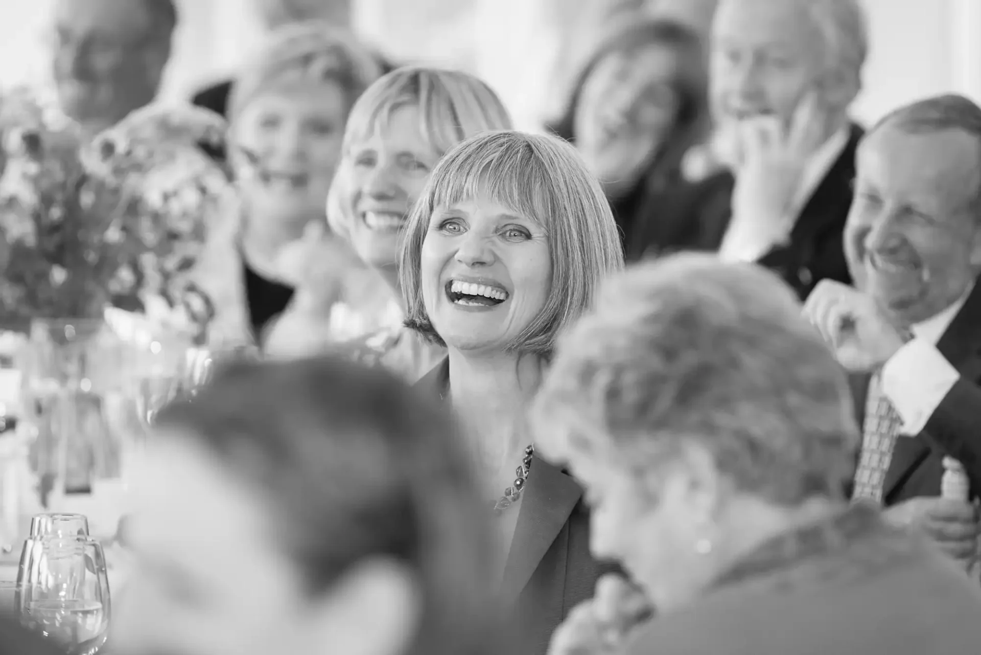 A black and white photo of a smiling woman with bobbed hair at a lively dinner event, surrounded by cheerful guests.
