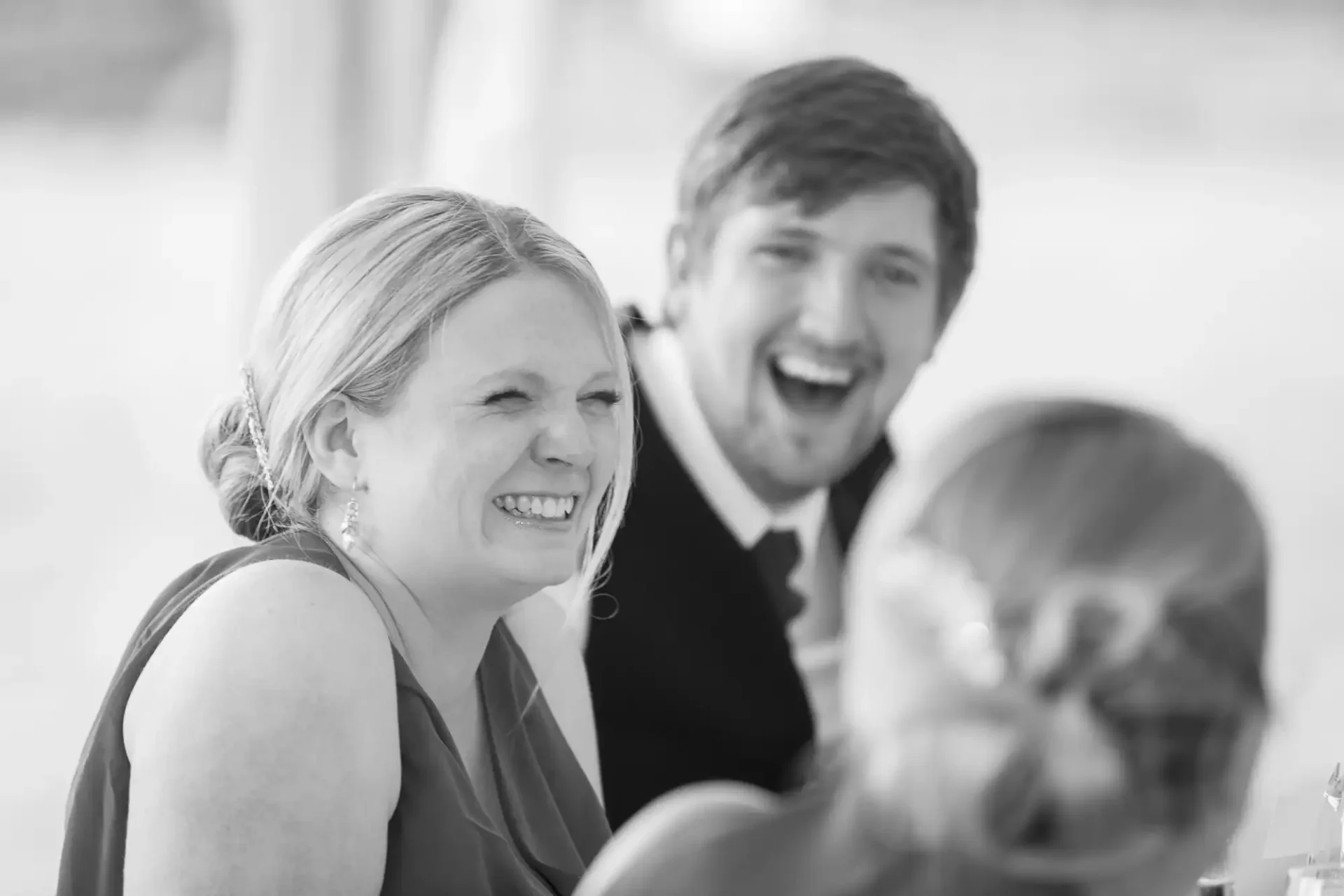 Two people laughing at a social event, captured in black and white, with a focus on a smiling woman and a man behind her.