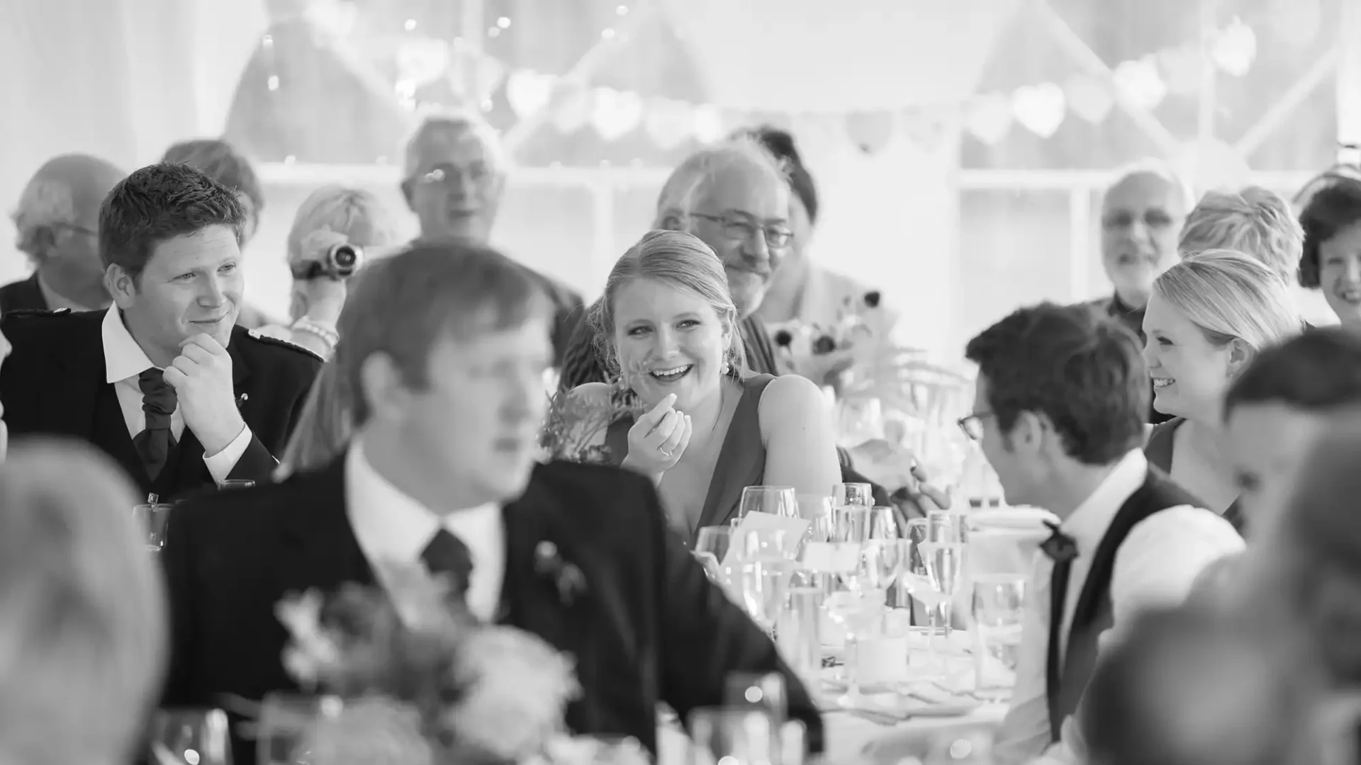 A black and white photo of people seated at a wedding reception, smiling and talking, with a festive atmosphere.