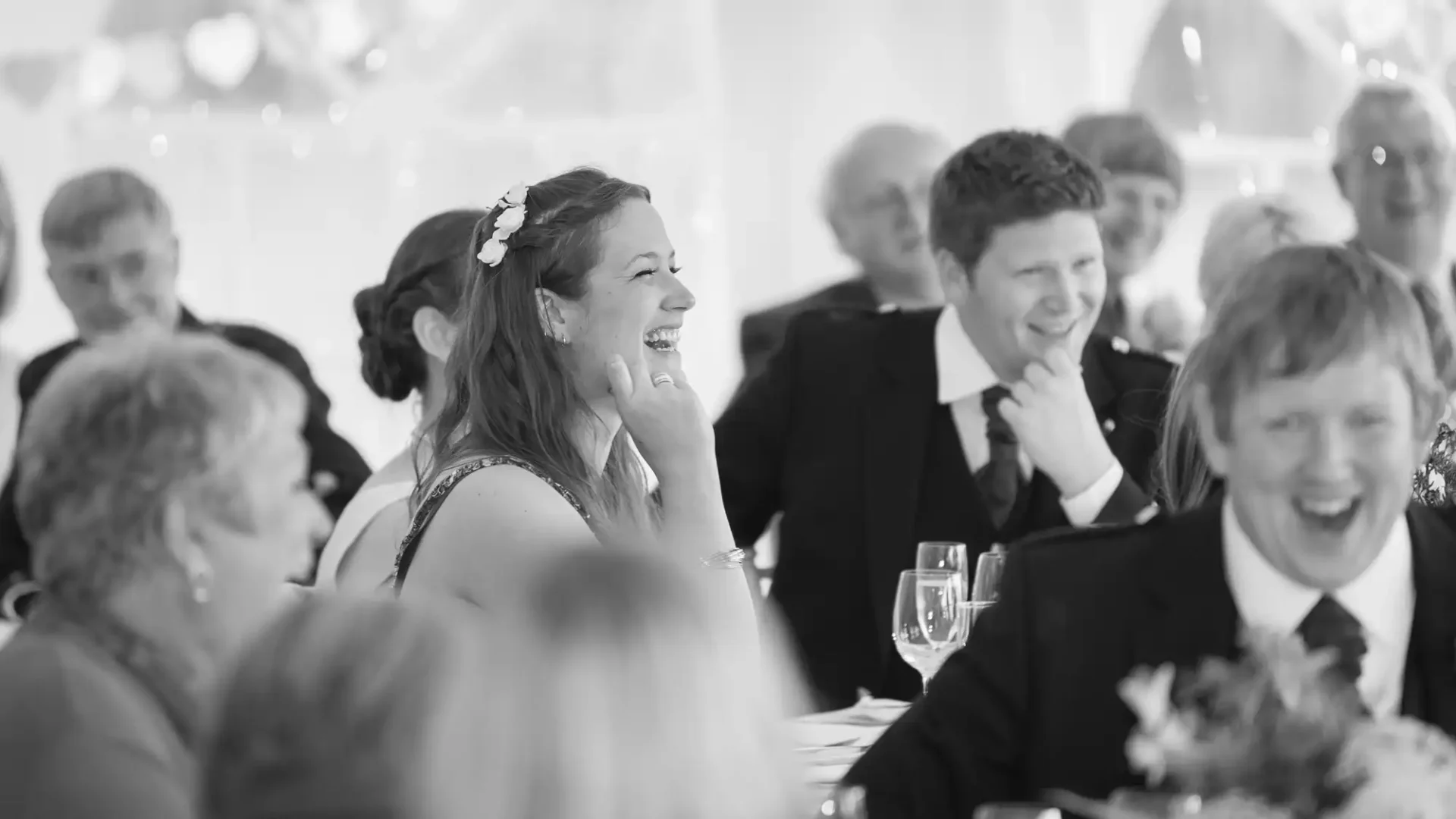 A black and white photo of a smiling woman with a floral hair accessory at a lively event, surrounded by guests laughing and conversing.