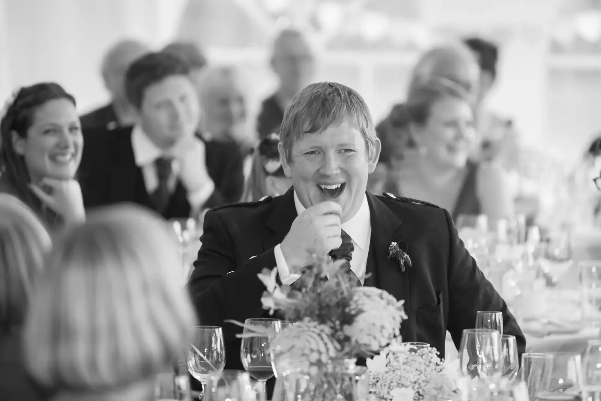 A man in a suit laughing at a wedding reception table in a black and white photograph, surrounded by other guests.