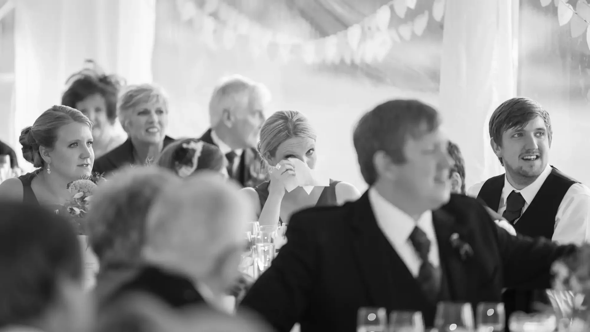 Black and white photo of guests sitting at a table during a wedding reception, capturing various expressions and interactions.