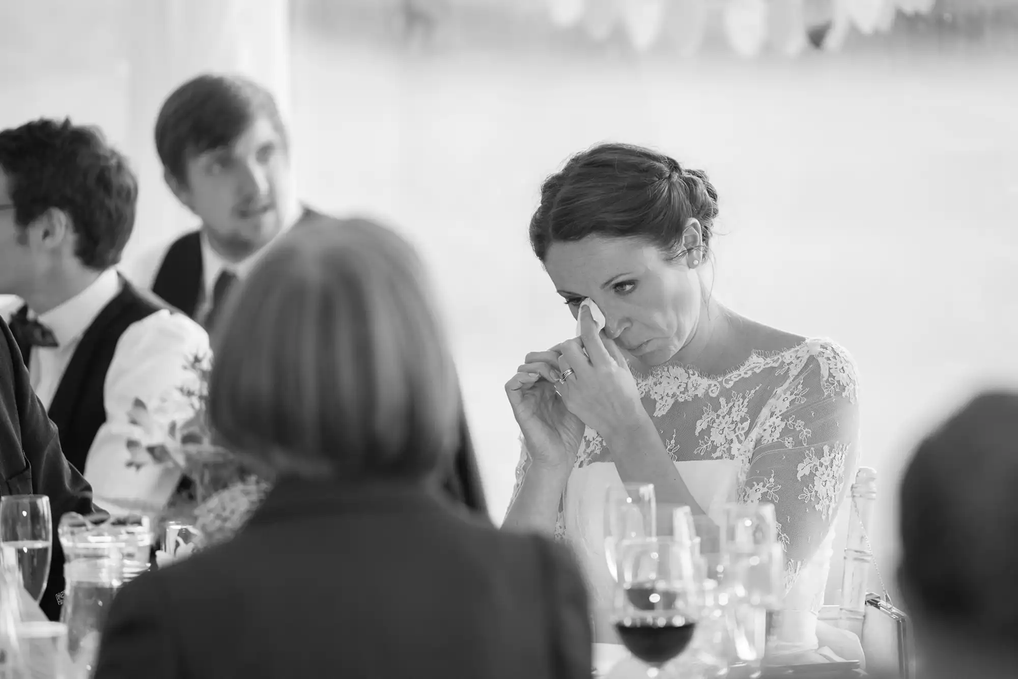 A bride in a lace dress wiping tears from her eyes at a wedding reception, with guests blurred in the background. black and white photo.