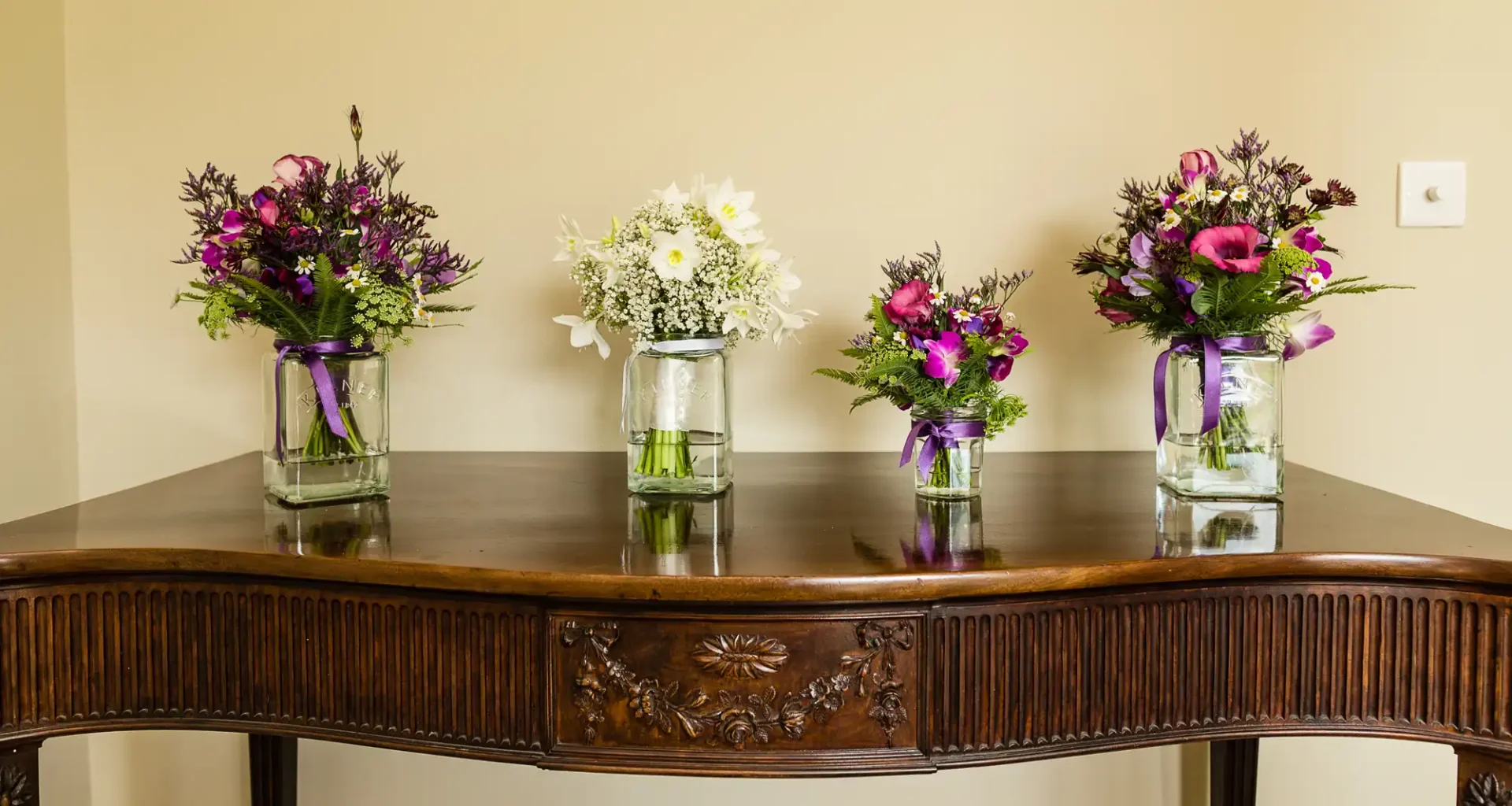Three vases with mixed flowers, two with purple blooms and one with white, placed on a wooden table against a beige wall.