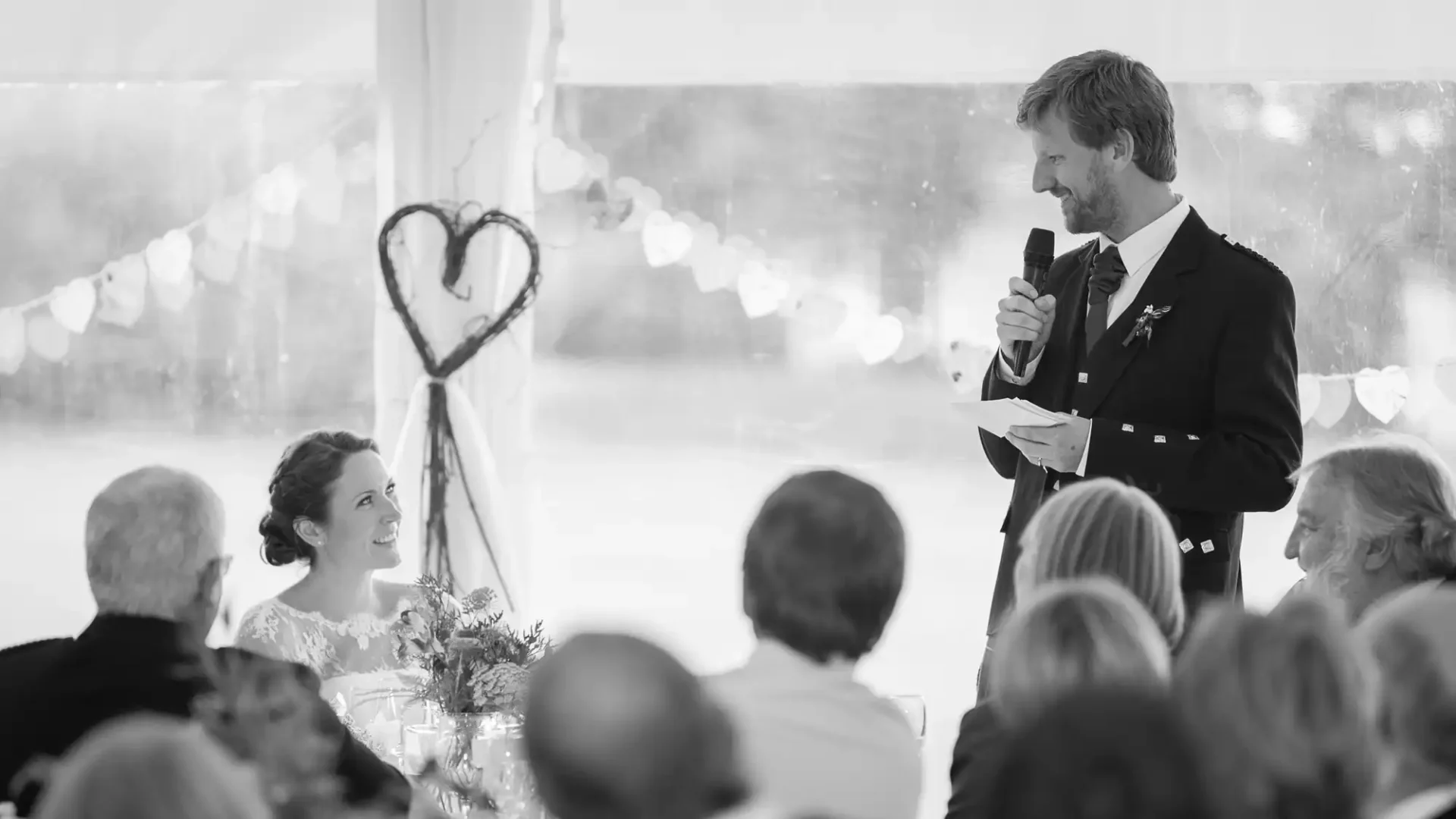 A man in a suit gives a speech at a wedding, holding a microphone and paper, facing a smiling bride seated among guests in a tent with string lights.