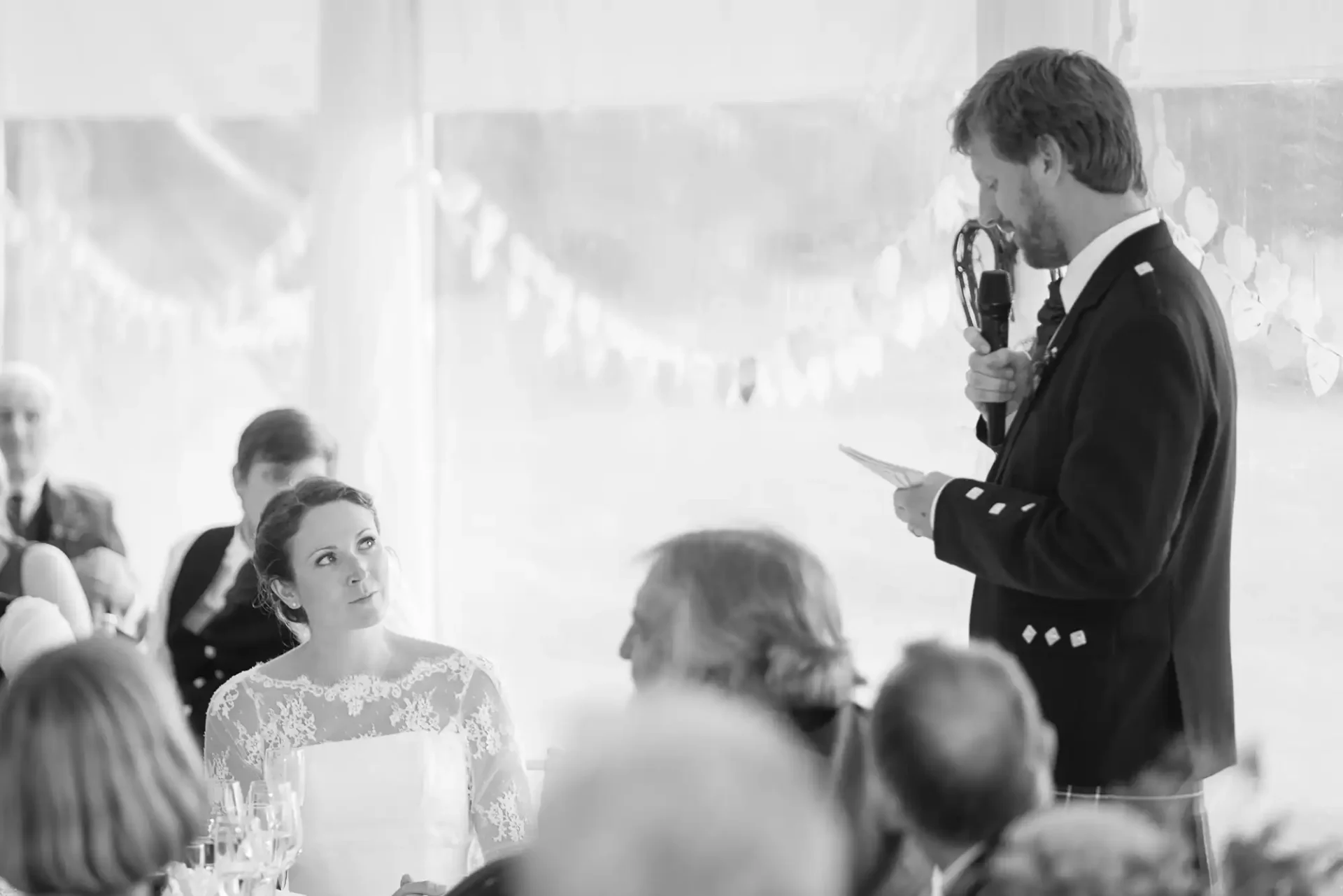 A man in a suit gives a speech with a microphone at a wedding reception, standing beside a seated bride, all in black and white.
