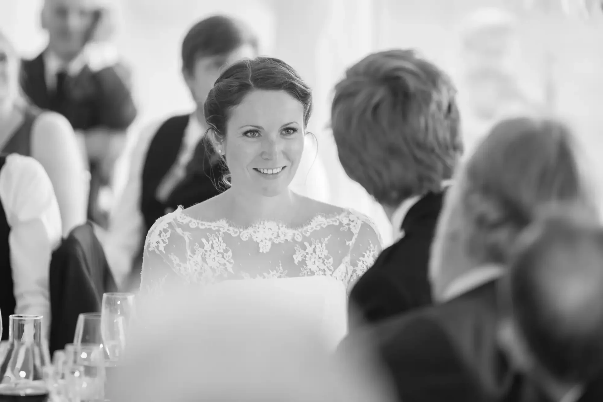 A bride in a lace dress smiling at a person across the table in a black-and-white photo of a wedding reception.