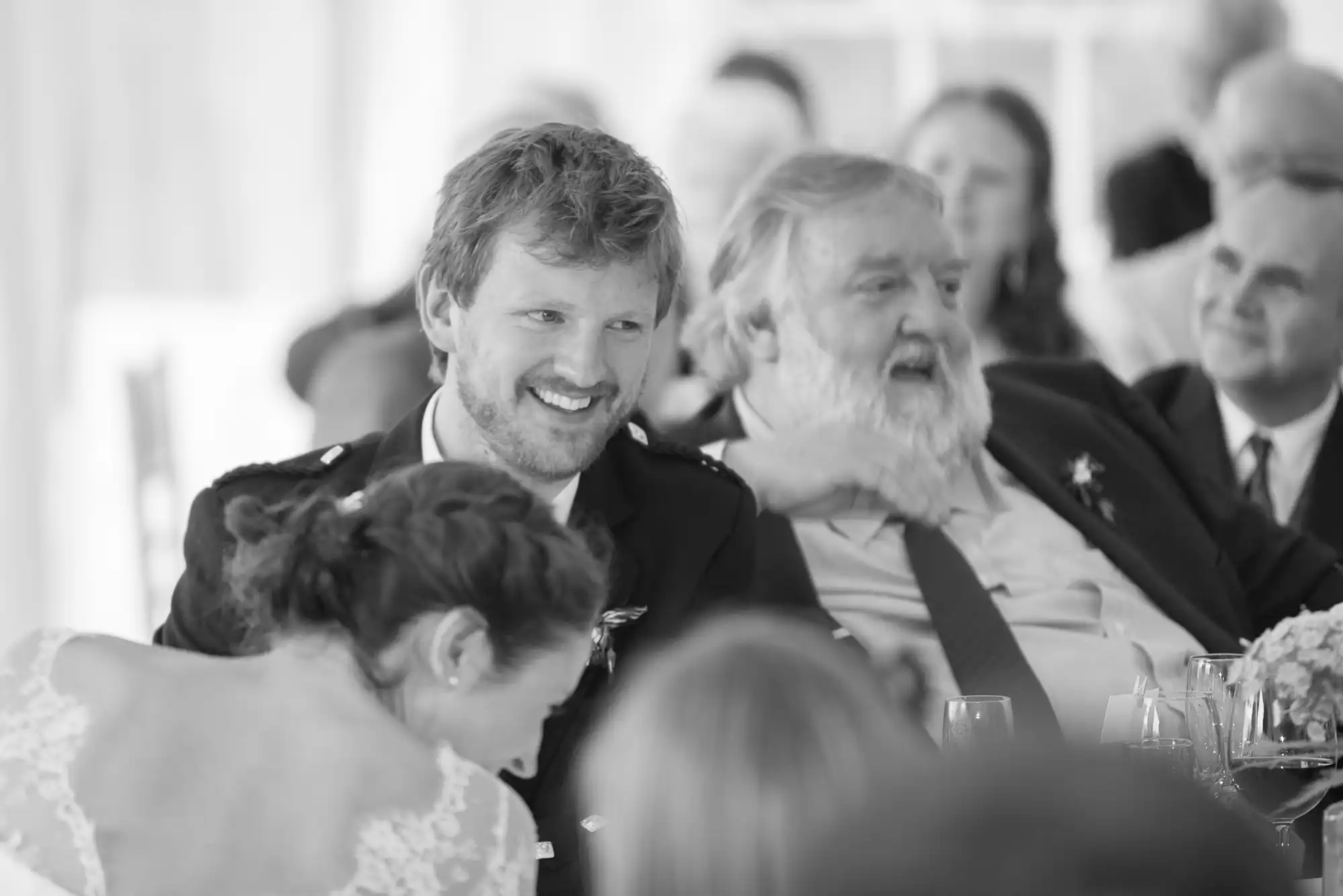 Black and white photo of two smiling men conversing at a wedding reception, with guests and floral decorations in the background.