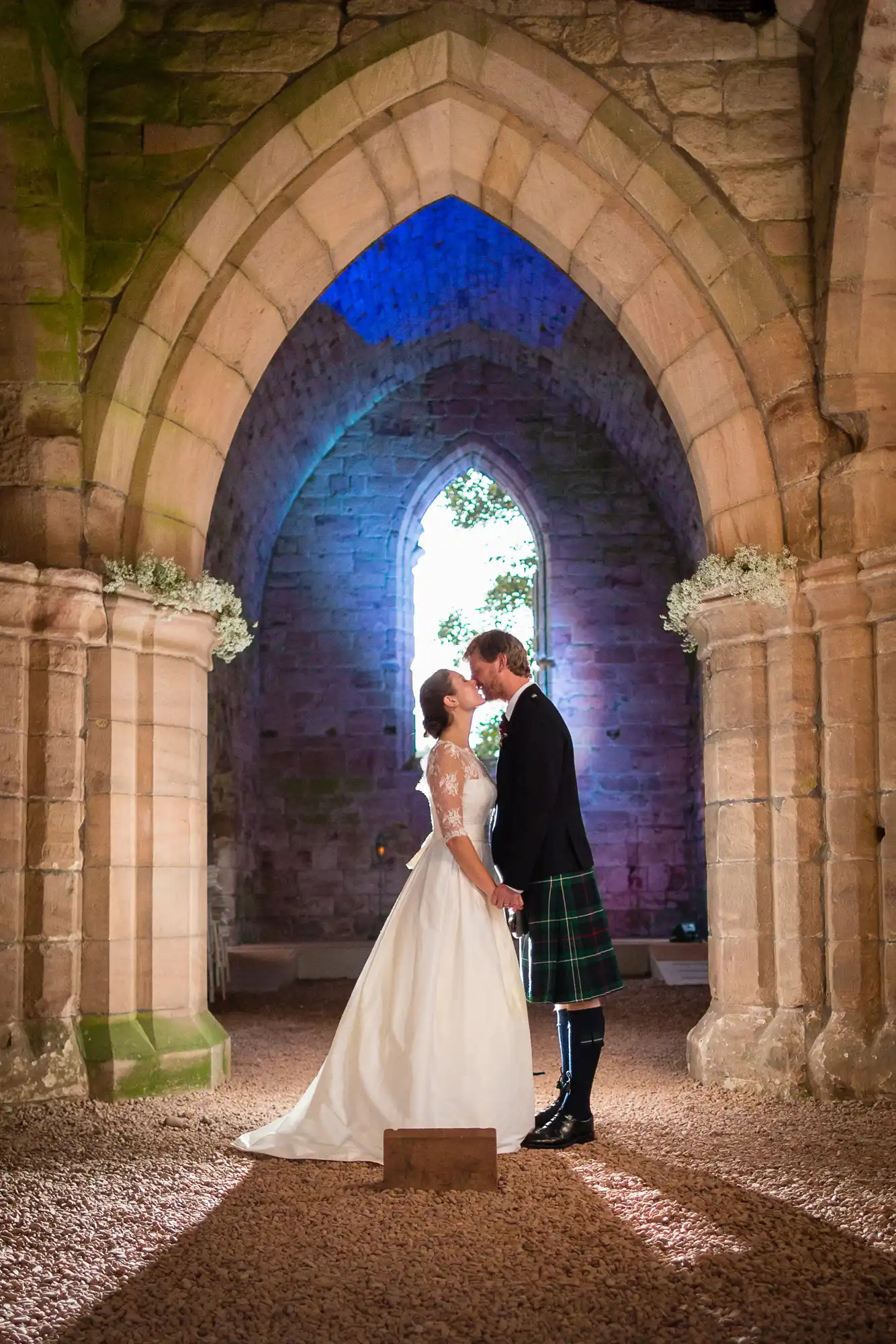 Bride and groom sharing a kiss inside a stone archway, the groom wearing a kilt, illuminated by warm lighting with a blue window in the background.