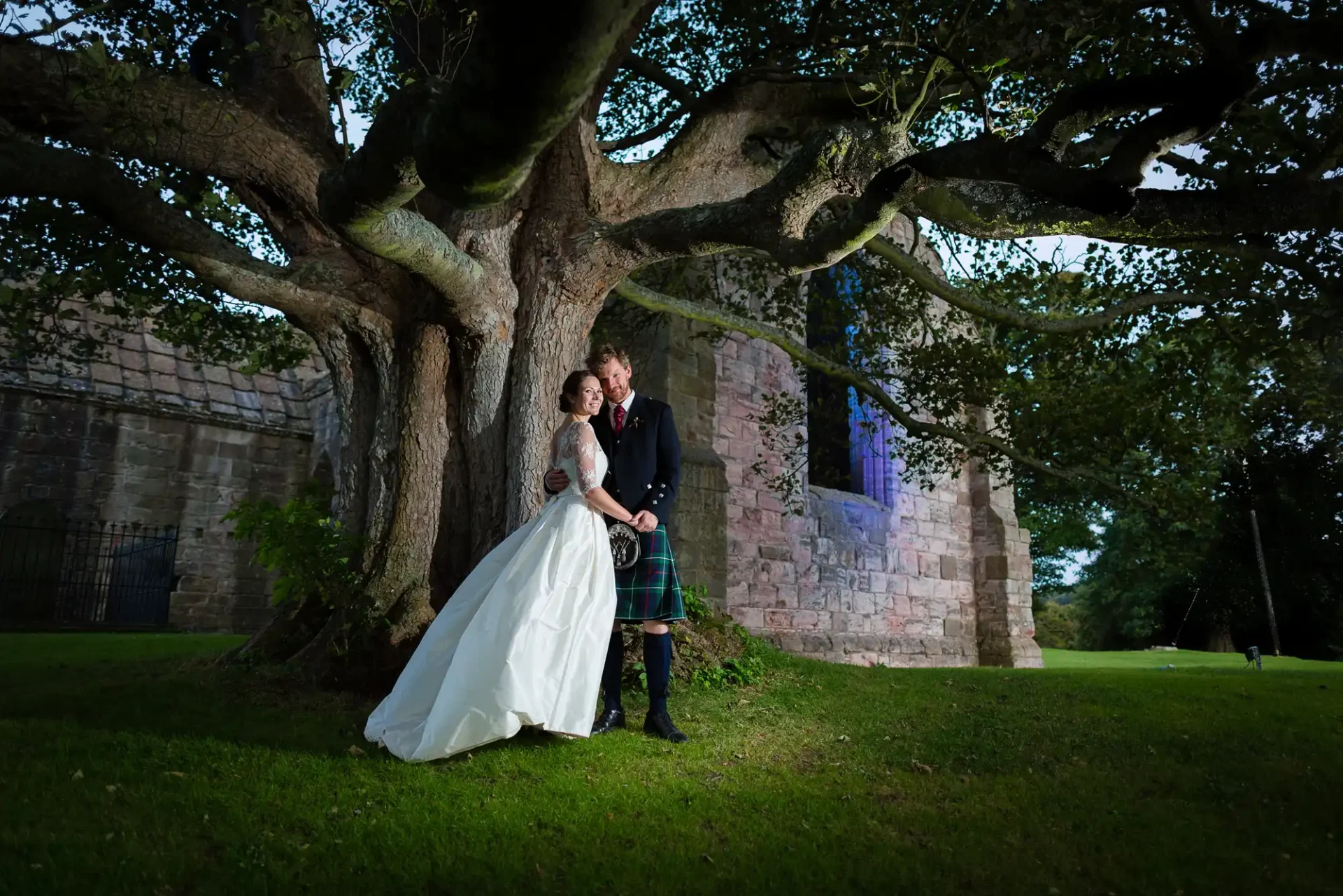A bride in a white dress and a groom in a kilt posing under a large tree near a stone building with illuminated windows.